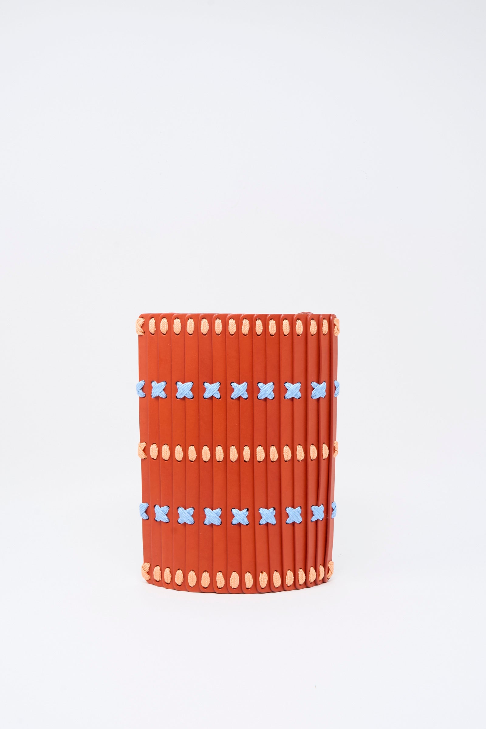 The Hatori Leather Crossbody 30 Artist Edition with Alex Dodge in Red is a cylindrical bag crafted from vegetable tanned leather and made in Japan. Features two horizontal rows of blue X-shaped accents, and small beige oval shapes along the top and bottom edges—demonstrating both artistry and meticulous craftsmanship.