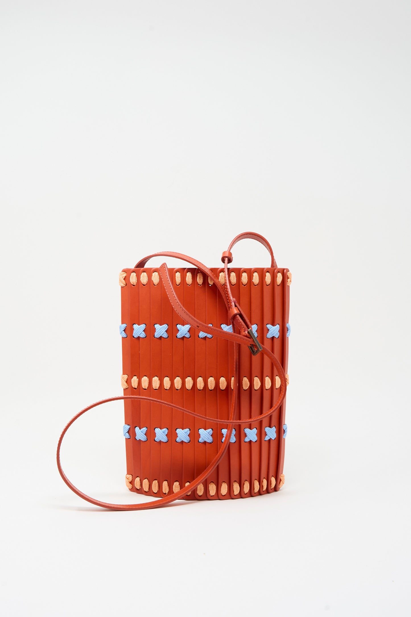 The Leather Crossbody 30 Artist Edition with Alex Dodge in Red by Hatori is a small cylindrical crossbody bag featuring vertical ridges, crafted from vegetable tanned leather. It is adorned with blue X patterns and beige oval accents. Made in Japan, this artist edition shoulder bag is displayed against a plain white background.