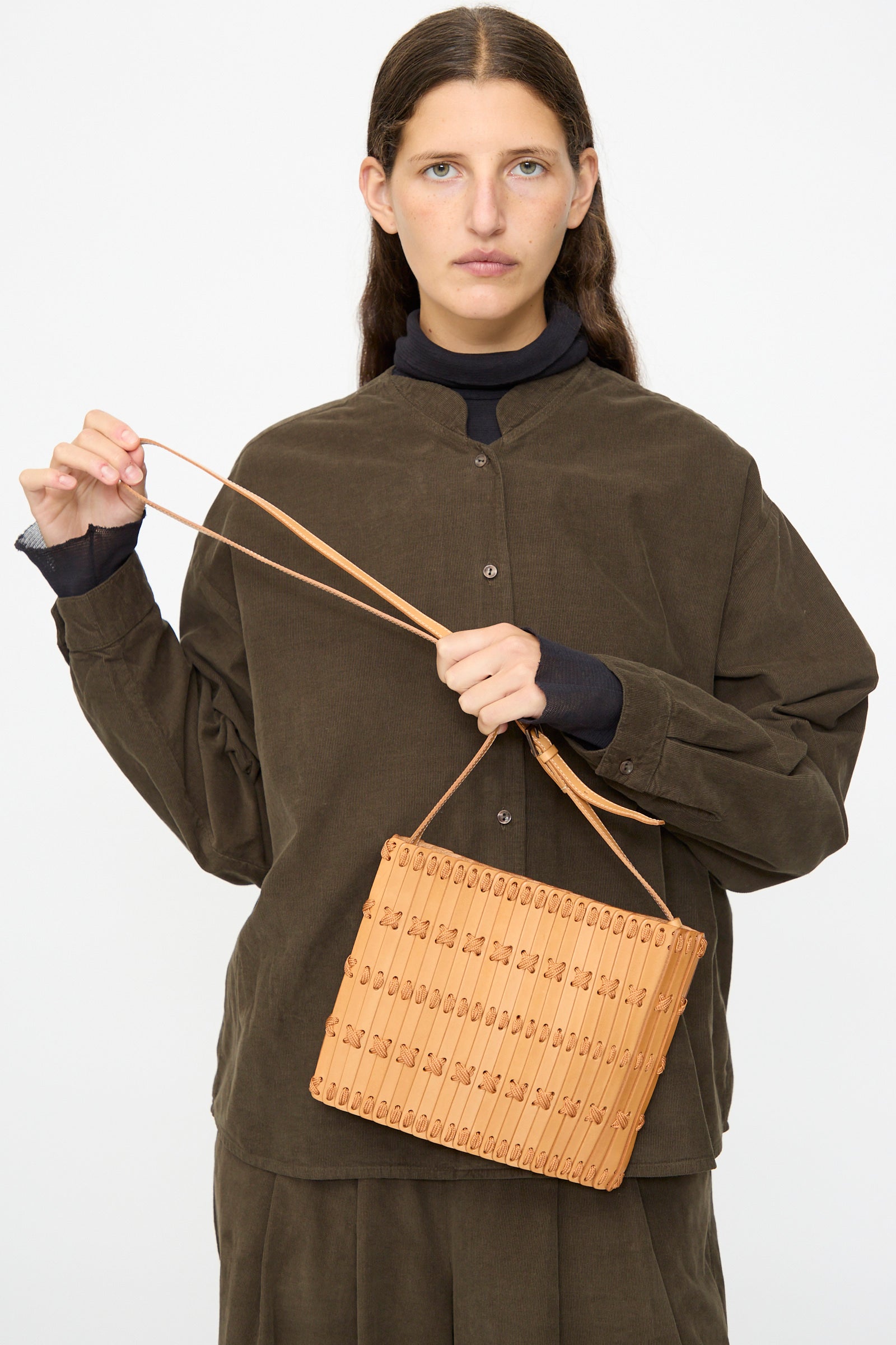 A person wearing a brown button-up shirt holds the Hatori Leather Crossbody 48 in Natural with Natural Lace by its strap against a plain background.