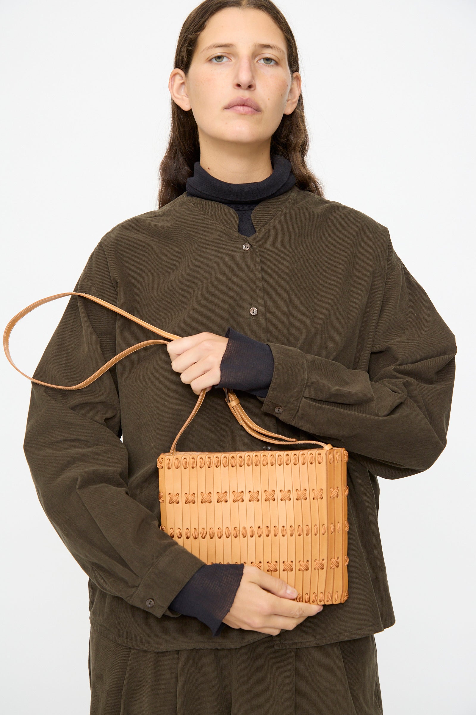 A person in a dark brown button-up shirt holds the Hatori Leather Crossbody 48 in Natural with Natural Lace, featuring a shoulder strap, against a plain background.