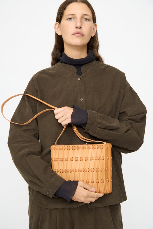 A person in a dark brown button-up shirt holds the Hatori Leather Crossbody 48 in Natural with Natural Lace, featuring a shoulder strap, against a plain background.