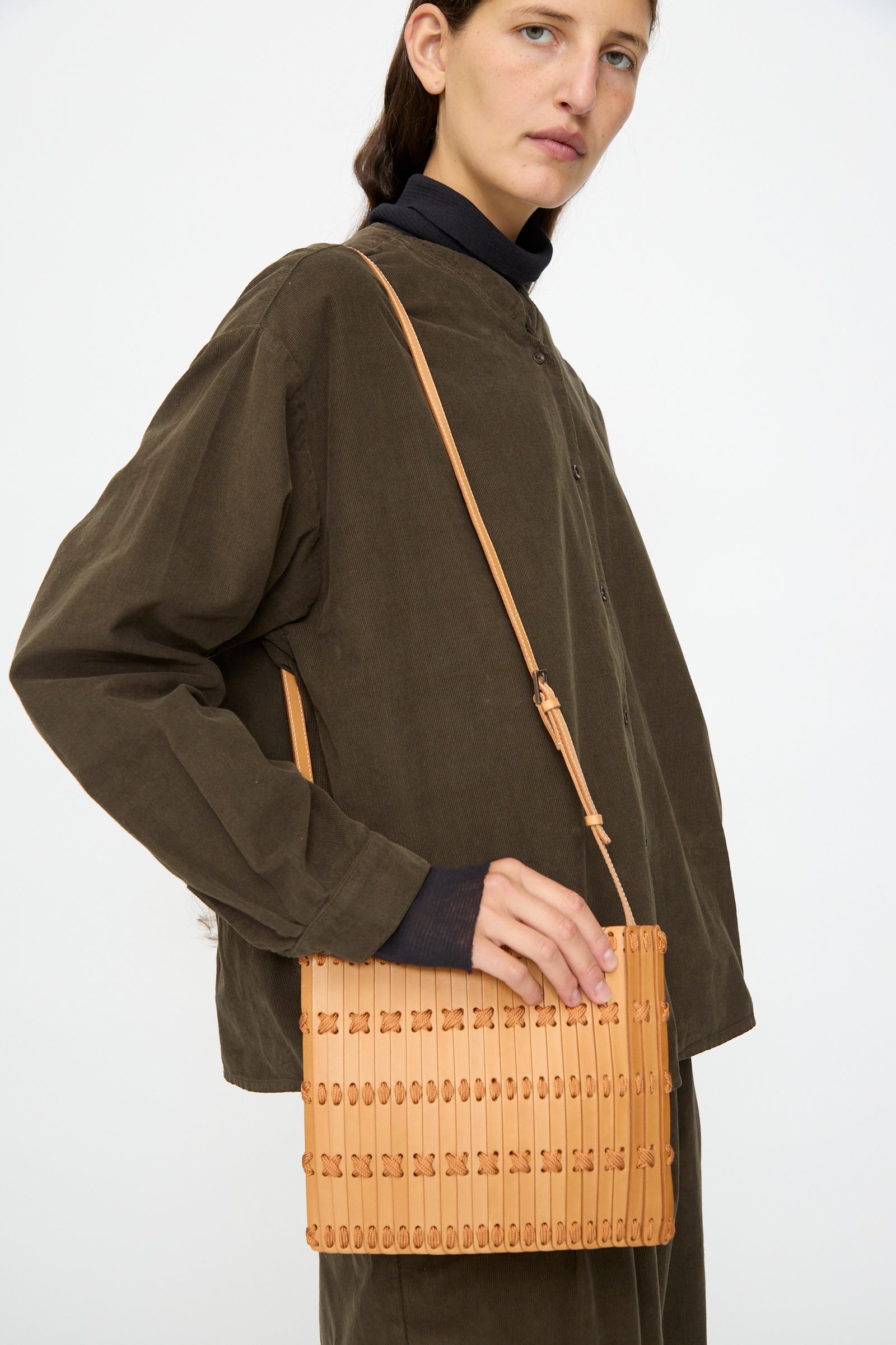 A person wearing a dark brown button-up shirt holds the Hatori Leather Crossbody 48 in Natural with Natural Lace, a handcrafted tan bag featuring an adjustable strap, against a plain background.