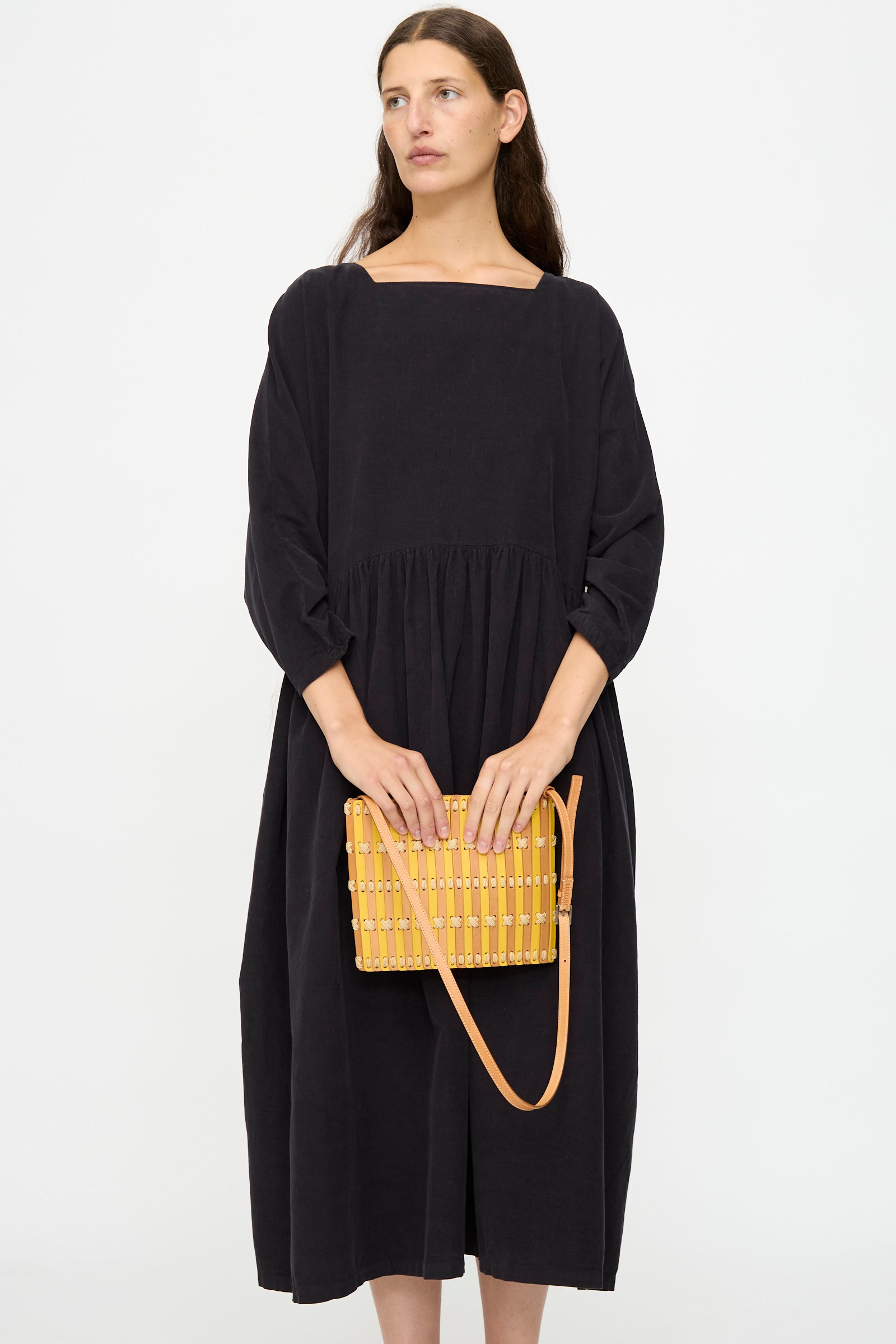 A woman in a loose black dress stands against a white background, holding Hatori's Leather Crossbody 48 in Natural and Yellow with Ecru Lace.
