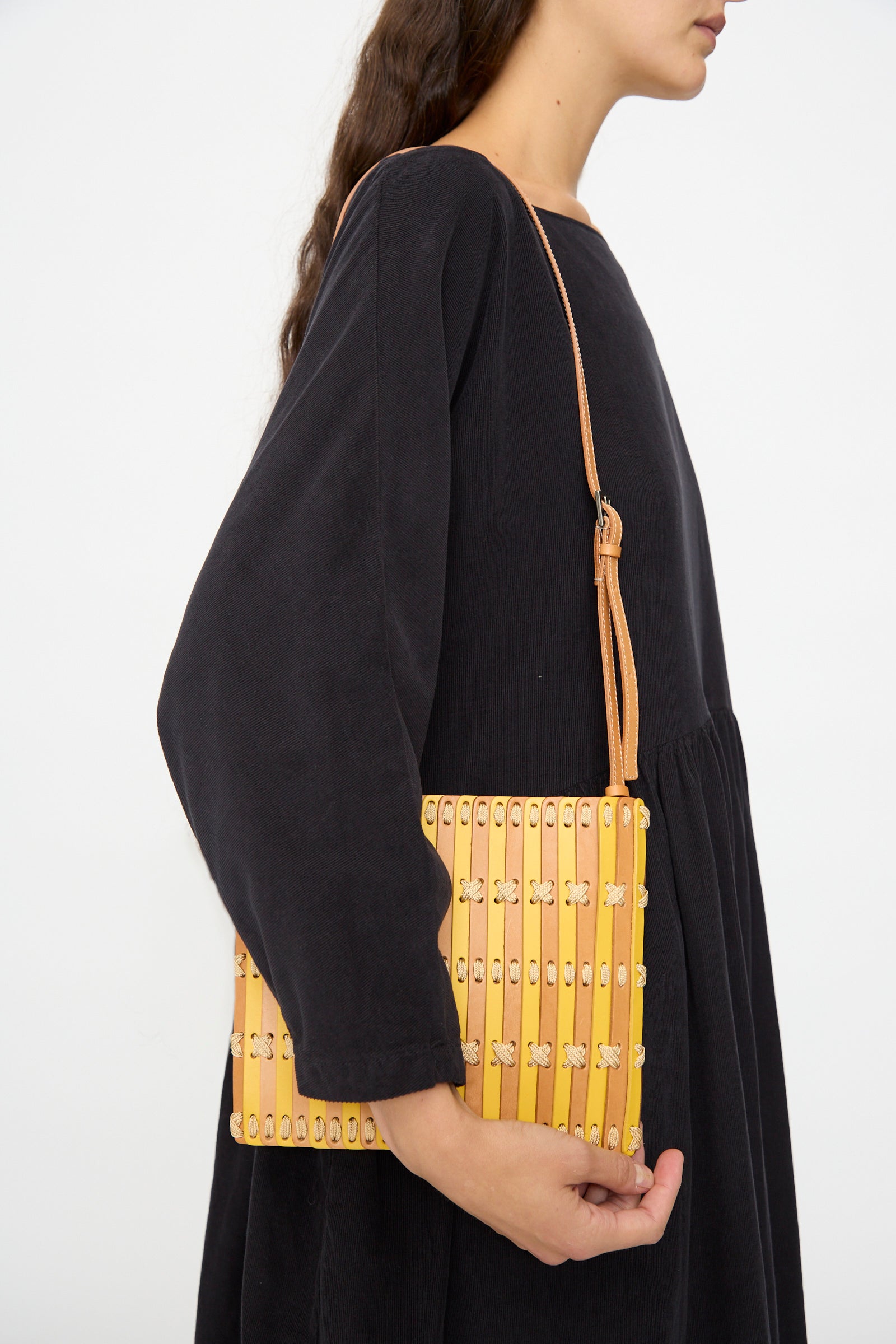 A woman in a black dress holds the Hatori Leather Crossbody 48 in Natural and Yellow with Ecru Lace, featuring handcrafted vertical stripes and metal accents, against a plain white background.