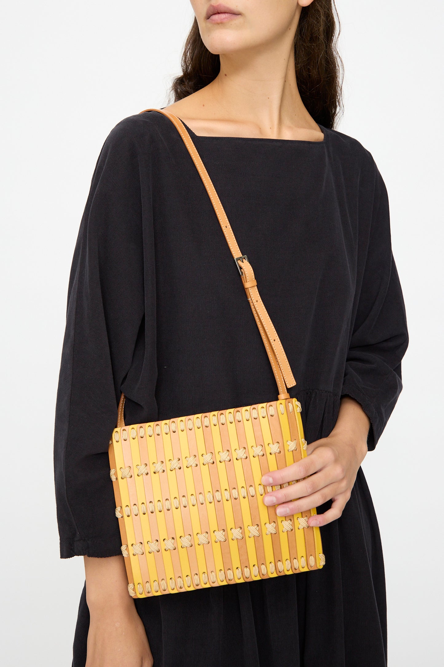 A woman in a black dress holds the Hatori Leather Crossbody 48 in Natural and Yellow with Ecru Lace, featuring a handcrafted slatted wooden design.