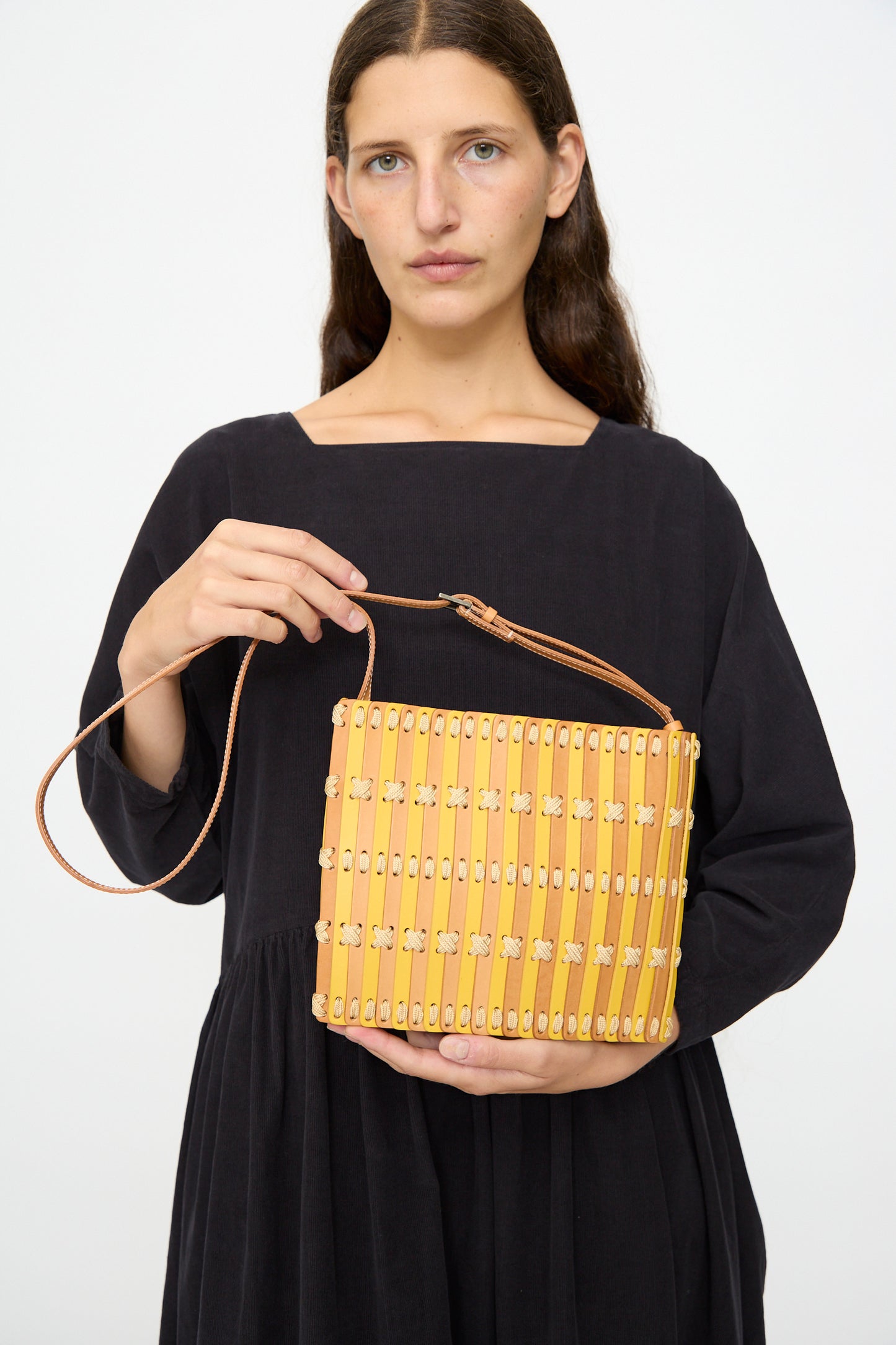 A woman in a black dress holds the Hatori Leather Crossbody 48 in Natural and Yellow with Ecru Lace, featuring a thin shoulder strap, against a plain background.
