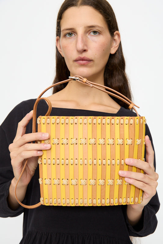 A woman in a black top holds the Hatori Leather Crossbody 48 in Natural and Yellow with Ecru Lace, featuring a vegetable tanned leather strap, positioned against a plain background.