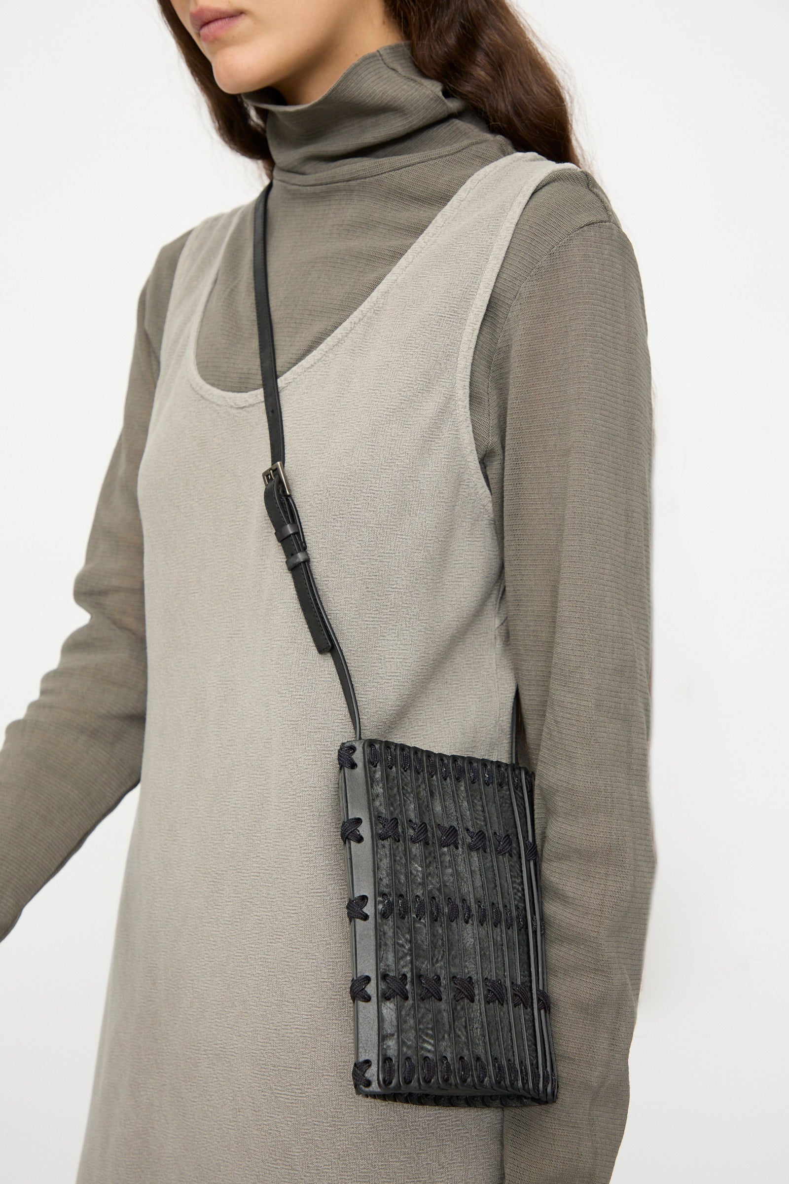 A woman in a taupe turtleneck dress with a sleeveless beige overlay carries the Hatori Leather Phone 26 Bag in Waxed Coated Black with Indigo Lace.