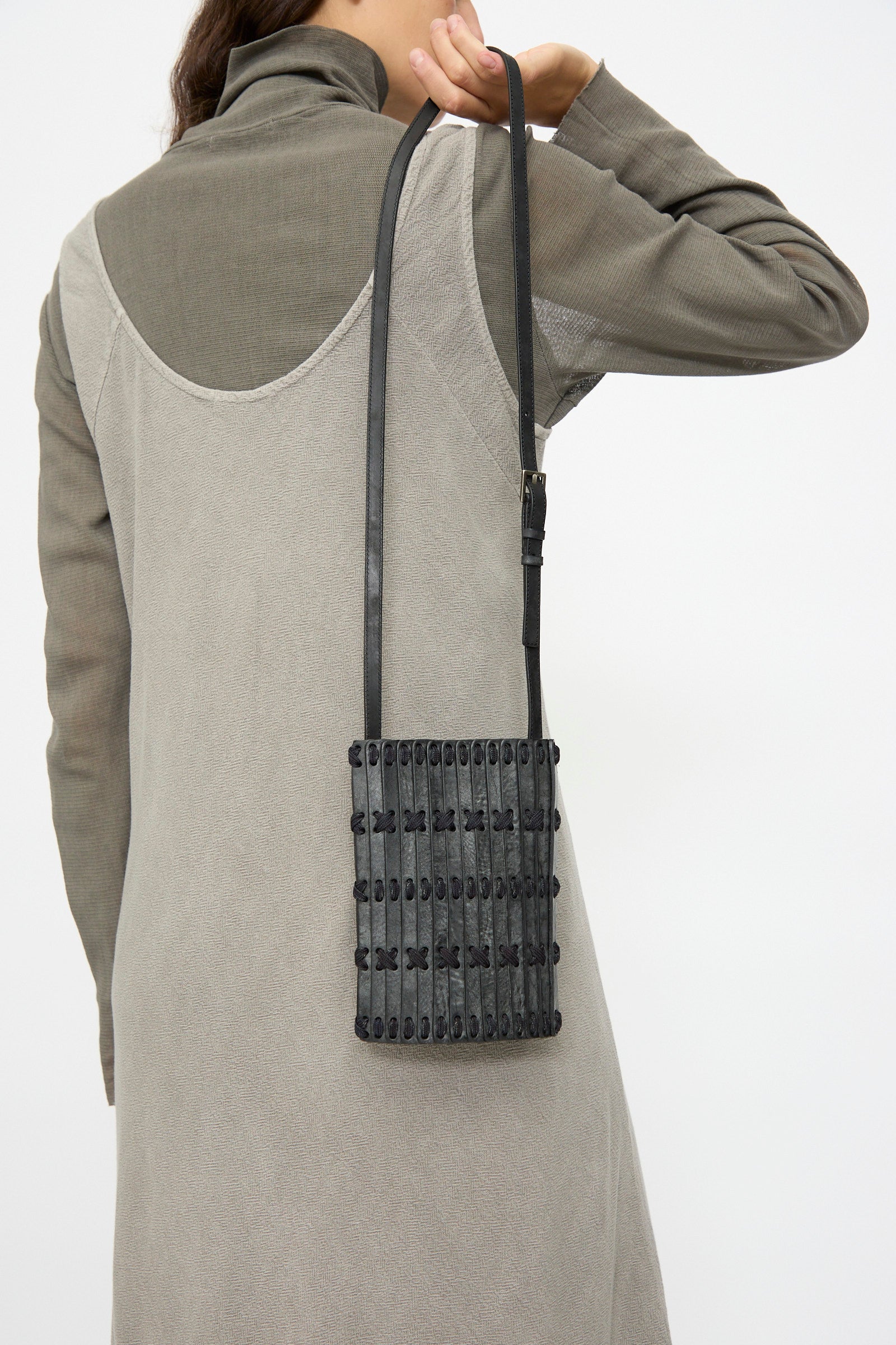 A person in a grey dress and long sleeves carries the Hatori Leather Phone 26 Bag in Waxed Coated Black with Indigo Lace, a small handcrafted rectangular bag made in Japan, over their shoulder.