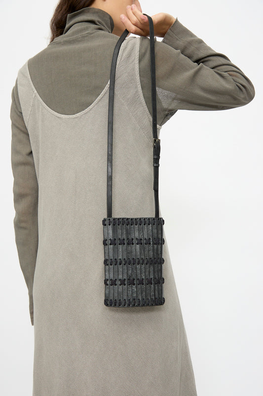 A person in a grey dress and long sleeves carries the Hatori Leather Phone 26 Bag in Waxed Coated Black with Indigo Lace, a small handcrafted rectangular bag made in Japan, over their shoulder.