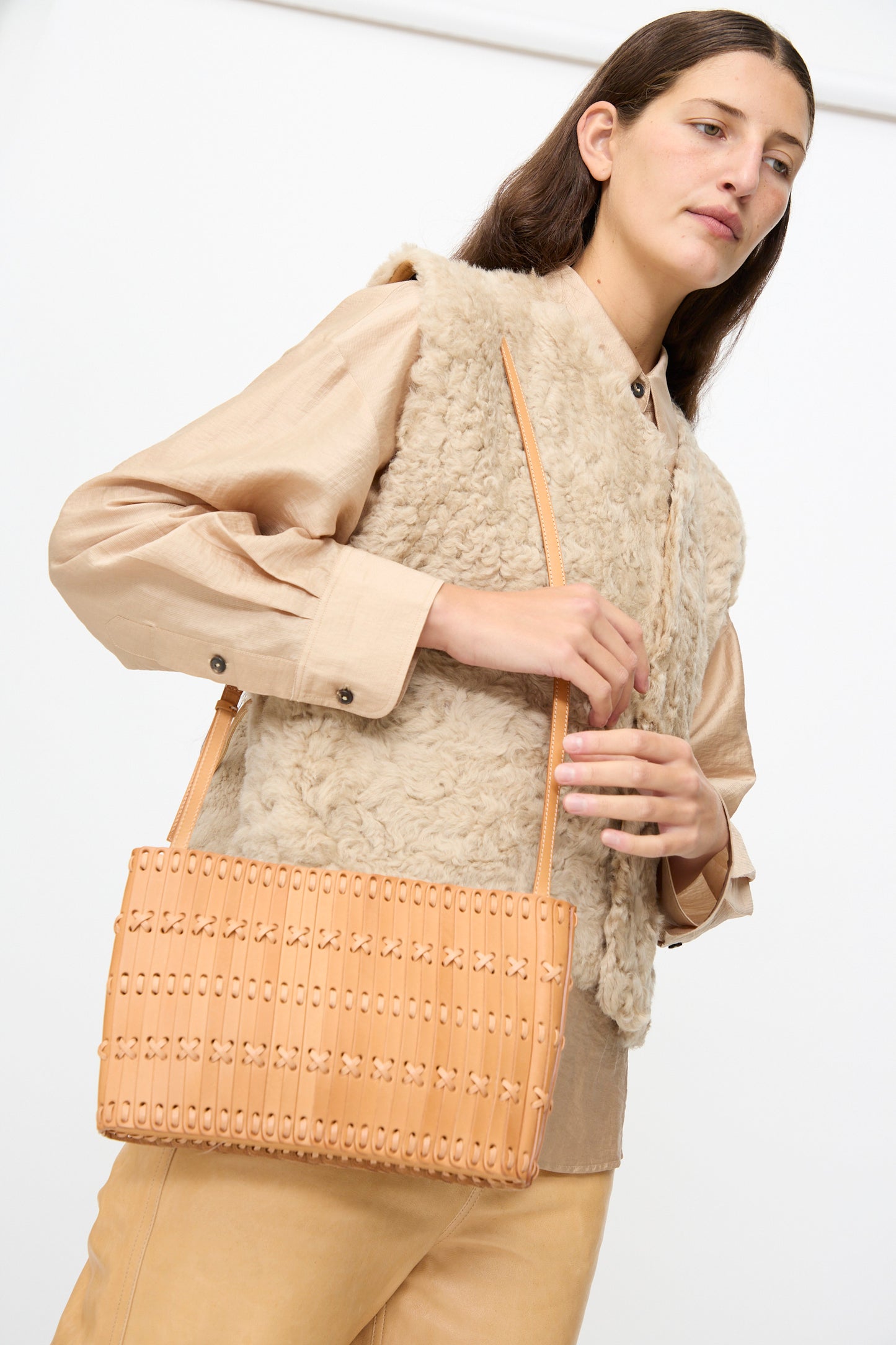 A woman wearing a beige shearling vest and tan pants holds the Hatori Leather Shoulder 60 Bag in Natural with Tan Lacing, a handcrafted Japanese shoulder bag made from vegetable tanned leather, against a plain background.