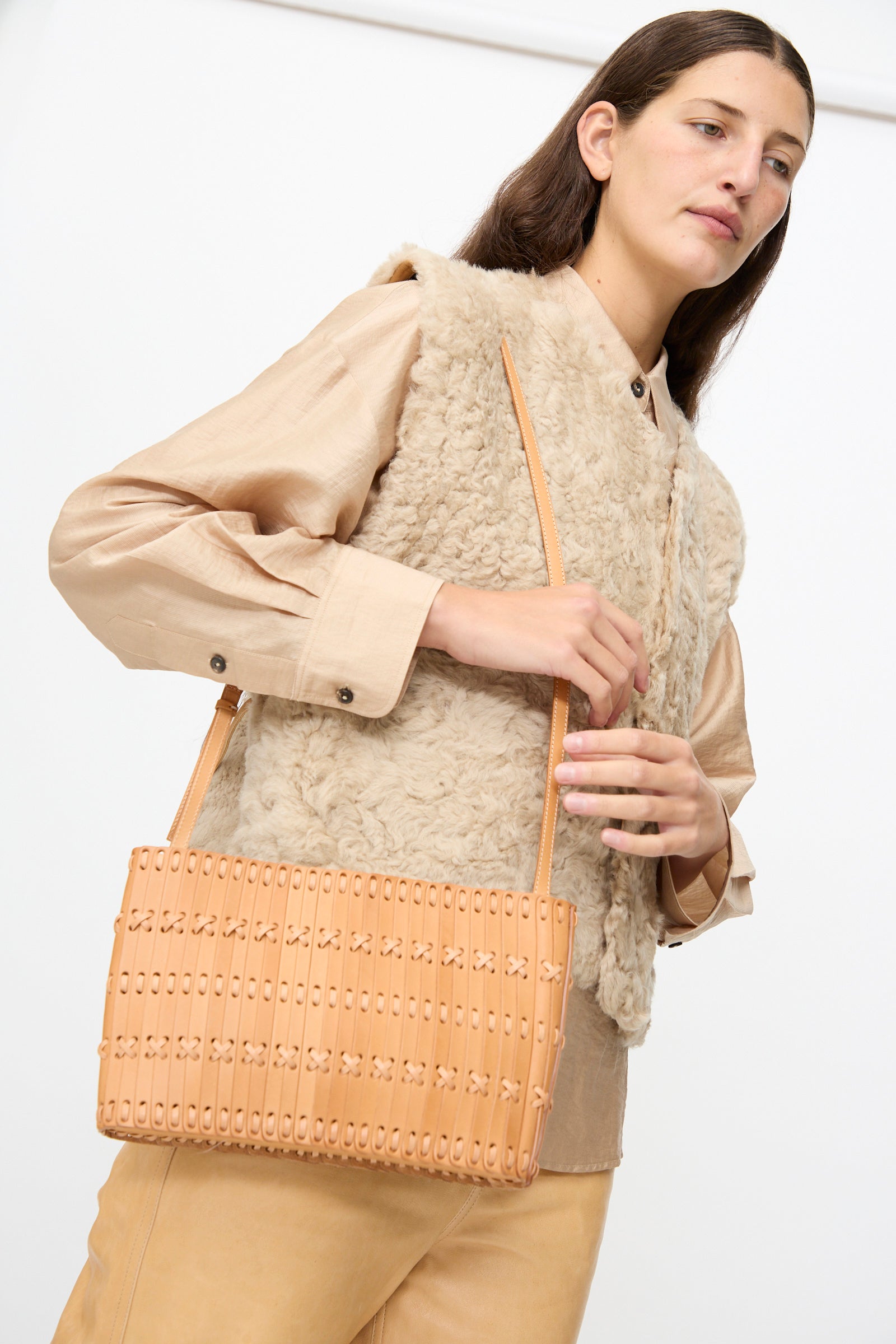 A woman wearing a beige shearling vest and tan pants holds the Hatori Leather Shoulder 60 Bag in Natural with Tan Lacing, a handcrafted Japanese shoulder bag made from vegetable tanned leather, against a plain background.
