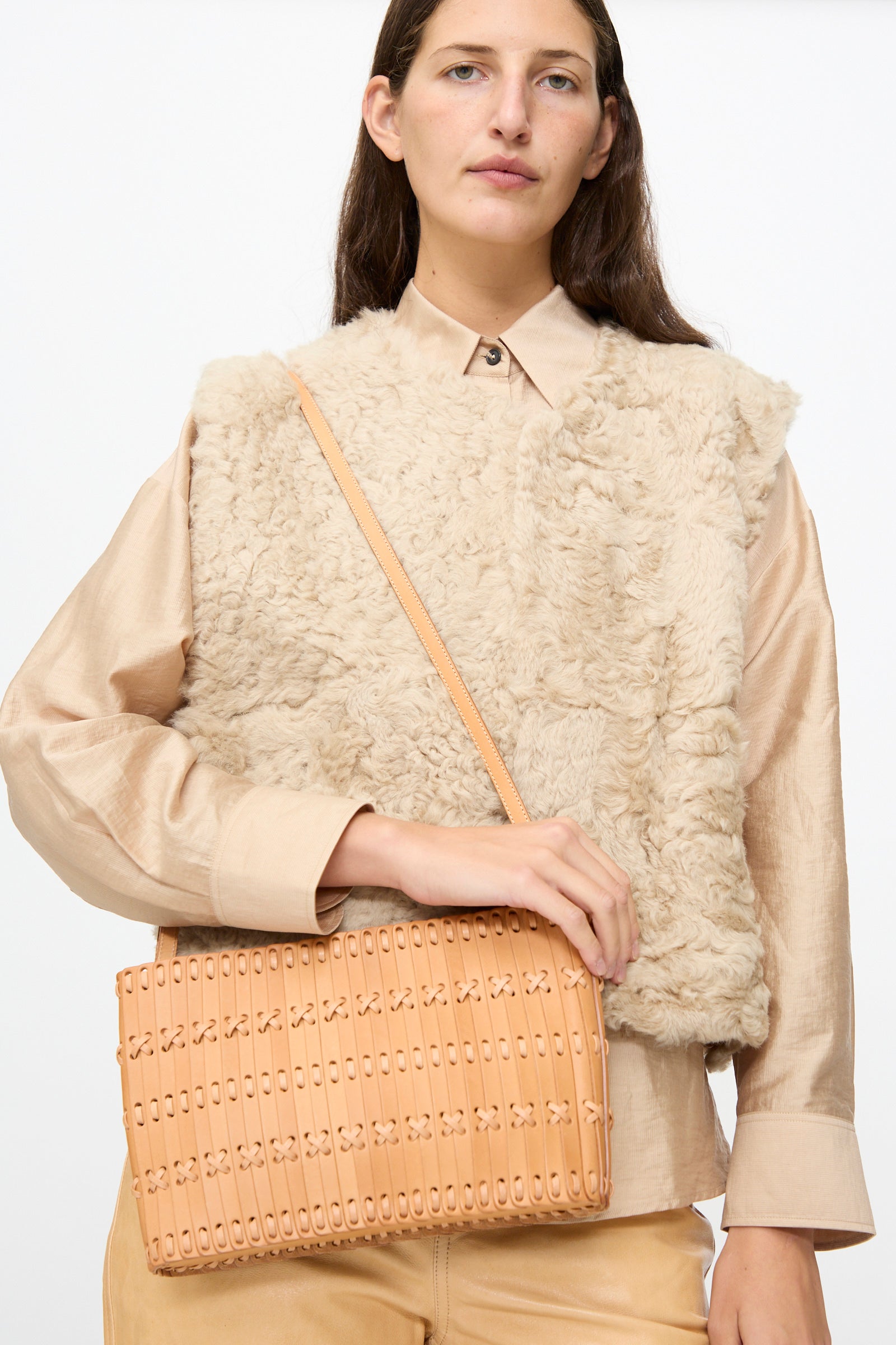 A woman wears a beige faux fur vest and holds the Hatori Leather Shoulder 60 Bag in Natural with Tan Lacing, a handcrafted vegetable-tanned leather shoulder bag featuring a patterned design, against a plain white background.