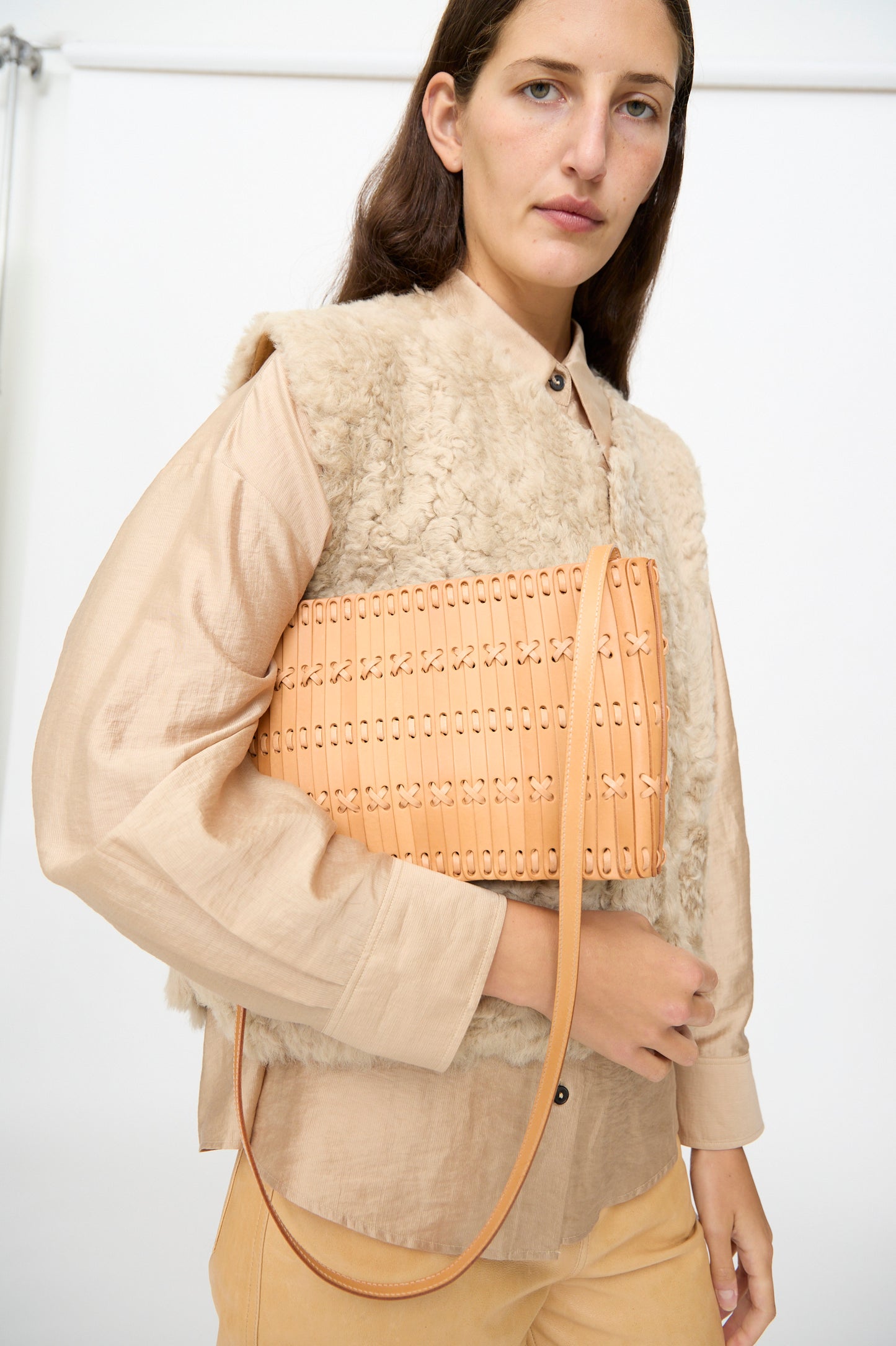 A woman wearing a beige shirt and shearling vest holds the Hatori Leather Shoulder 60 Bag in Natural with Tan Lacing, a handcrafted piece, standing against a plain background.