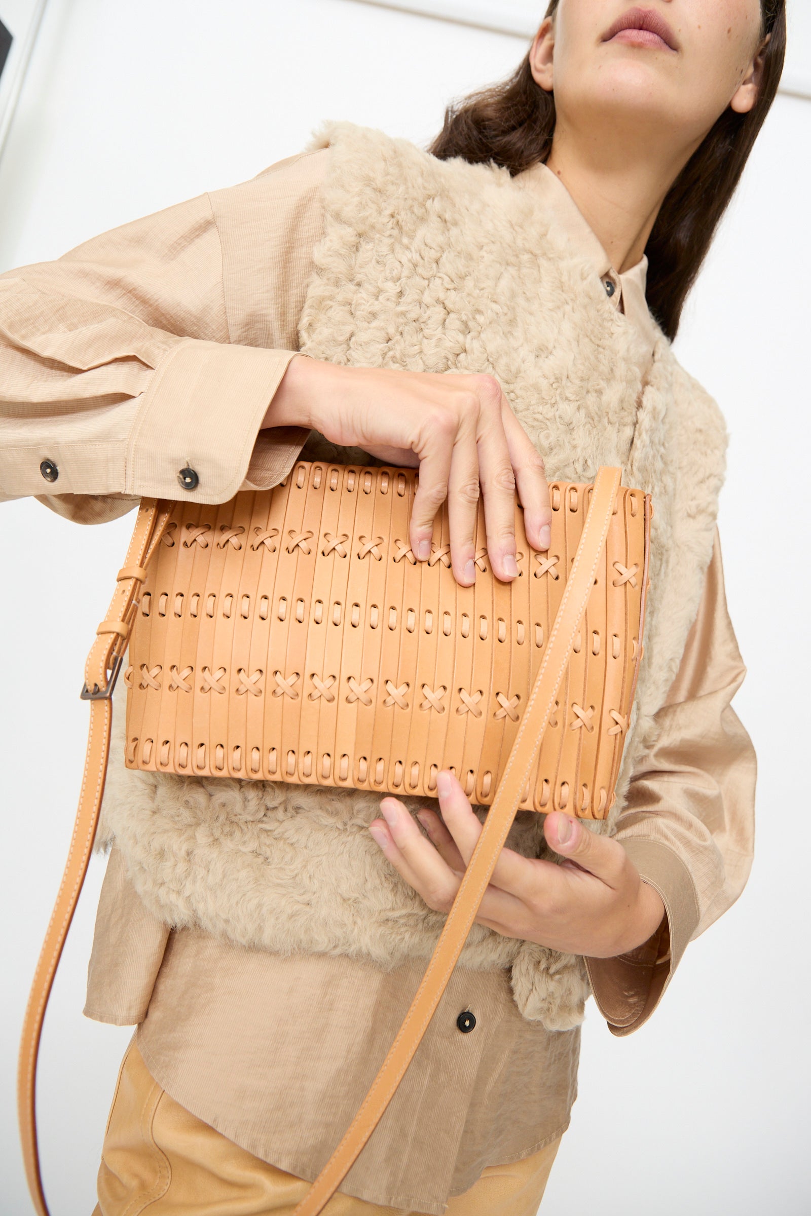 A person wearing a beige shirt and shearling vest holds the Hatori Leather Shoulder 60 Bag in Natural with Tan Lacing—a handcrafted crossbody featuring a patterned design in vegetable tanned leather—while standing indoors.
