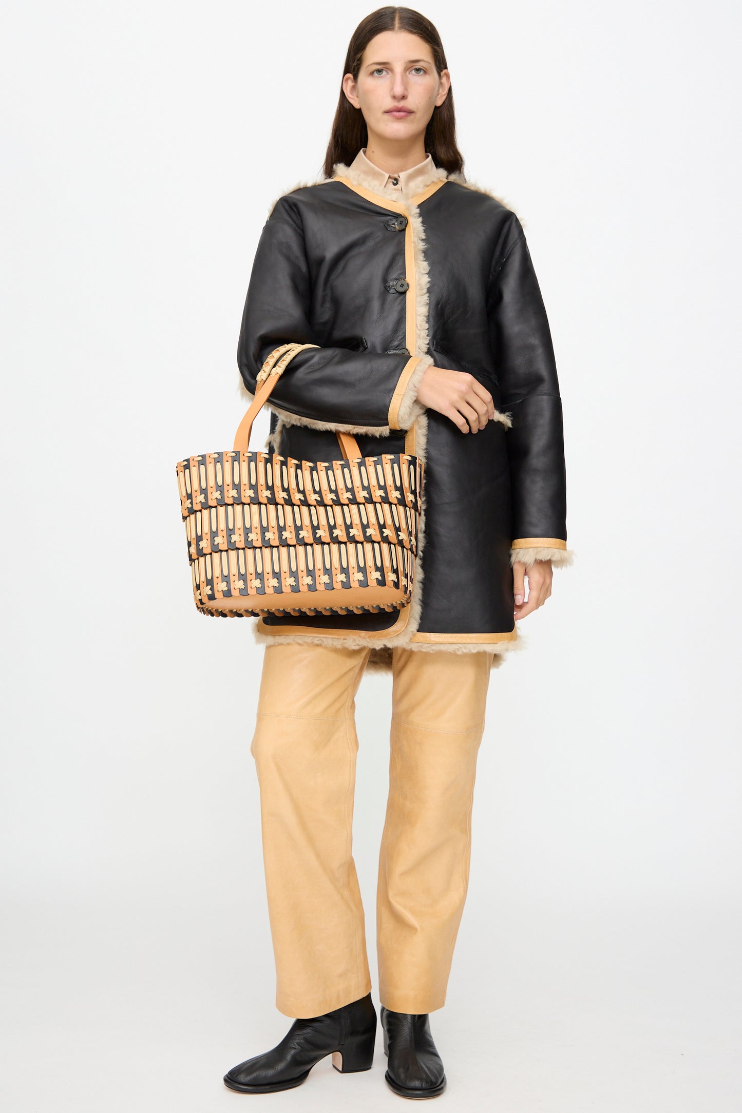 A person stands facing forward against a plain white background, wearing a black shearling coat, tan pants, and black boots, holding the Hatori Leather Tote 198 in Bi-Color with Tan Lacing—a large handcrafted leather bag made in Japan.