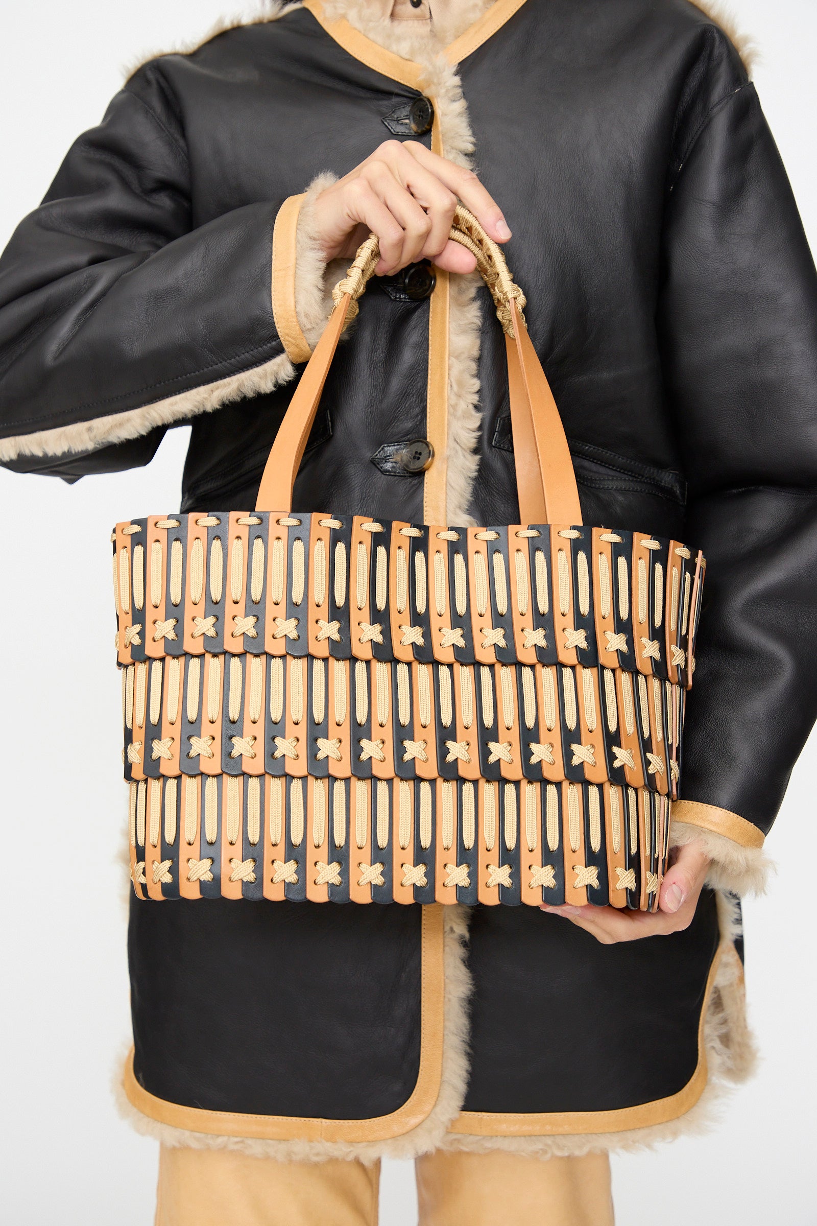 A person wearing a black and tan shearling coat holds the Hatori Leather Tote 198 in Bi-Color with Tan Lacing, a handcrafted bag featuring a geometric tan and black pattern and tan handles, made in Japan.
