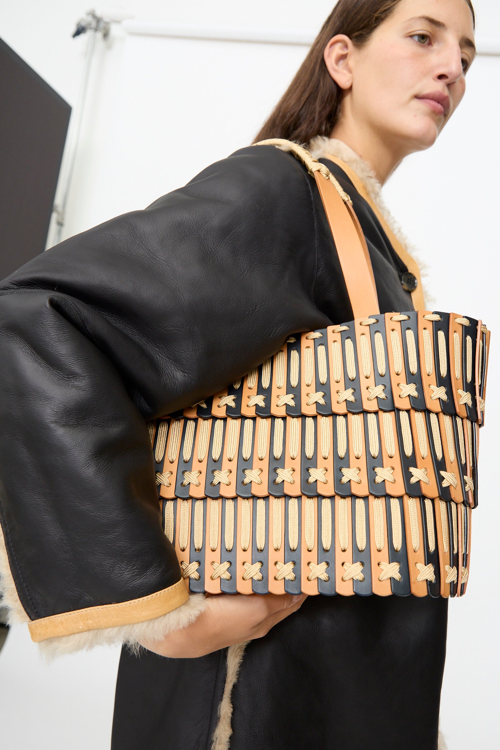 A person wearing a black shearling coat holds the Hatori Leather Tote 198 in Bi-Color with Tan Lacing—a handcrafted rectangular leather handbag featuring tan and black patterns—against a plain indoor background.