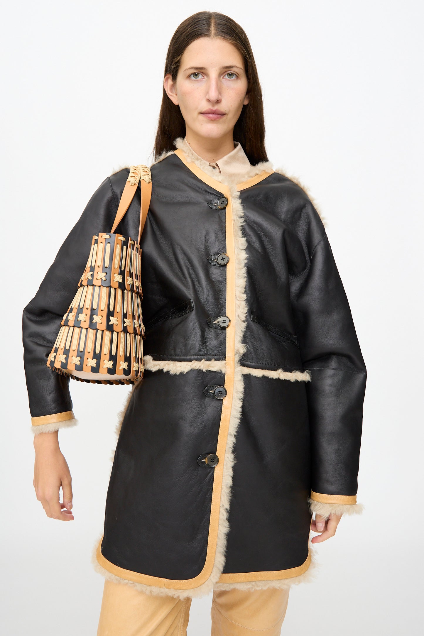 A woman wearing a black shearling coat with tan trim stands against a plain white background, carrying the Hatori Leather Tote 198 in Bi-Color with Tan Lacing over her shoulder.