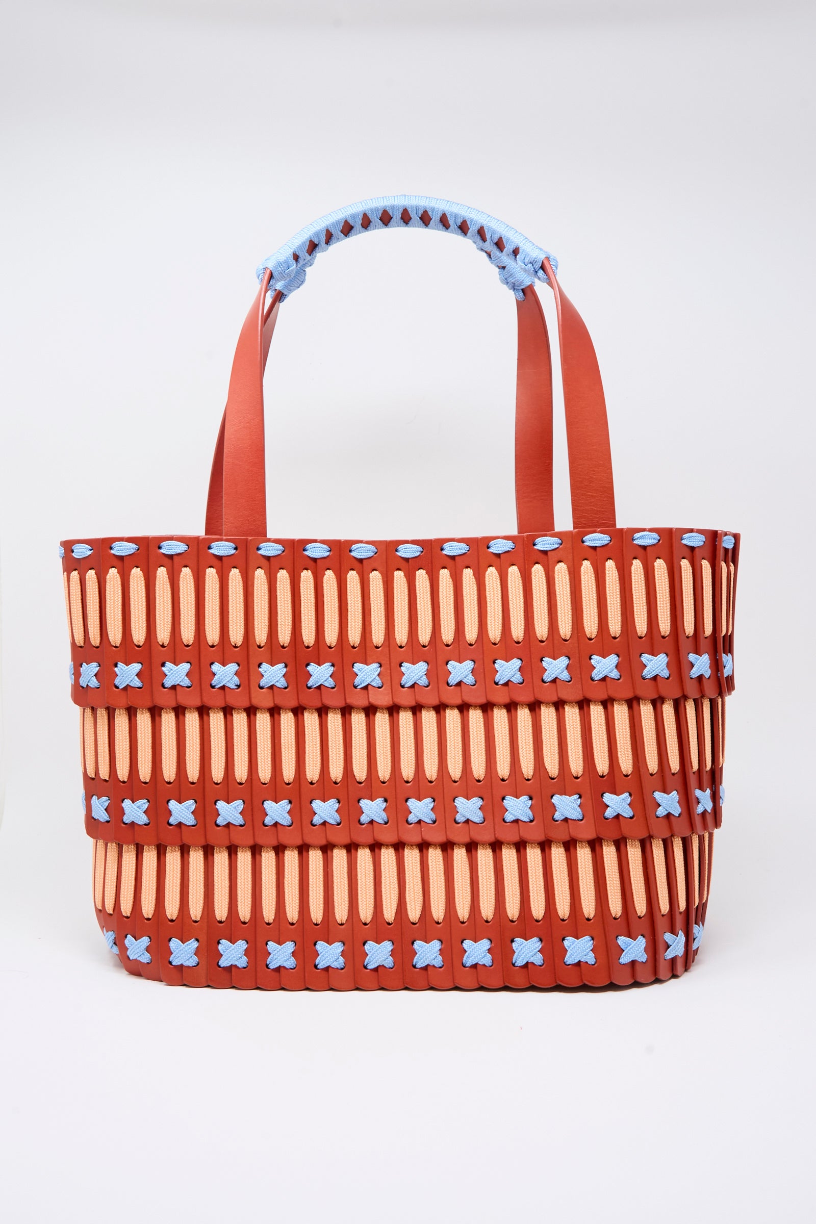 Introducing the Hatori Leather Tote 198 Artist Edition with Alex Dodge in Red—a handcrafted leather tote meticulously constructed from premium vegetable tanned leather. Featuring a vibrant palette of red, tan, and blue, this woven masterpiece showcases vertical tan strips and bold blue X-shaped accents. Two red handles with intricate blue weaving at the top add to its artistic flair, blending exceptional craftsmanship with functional design.