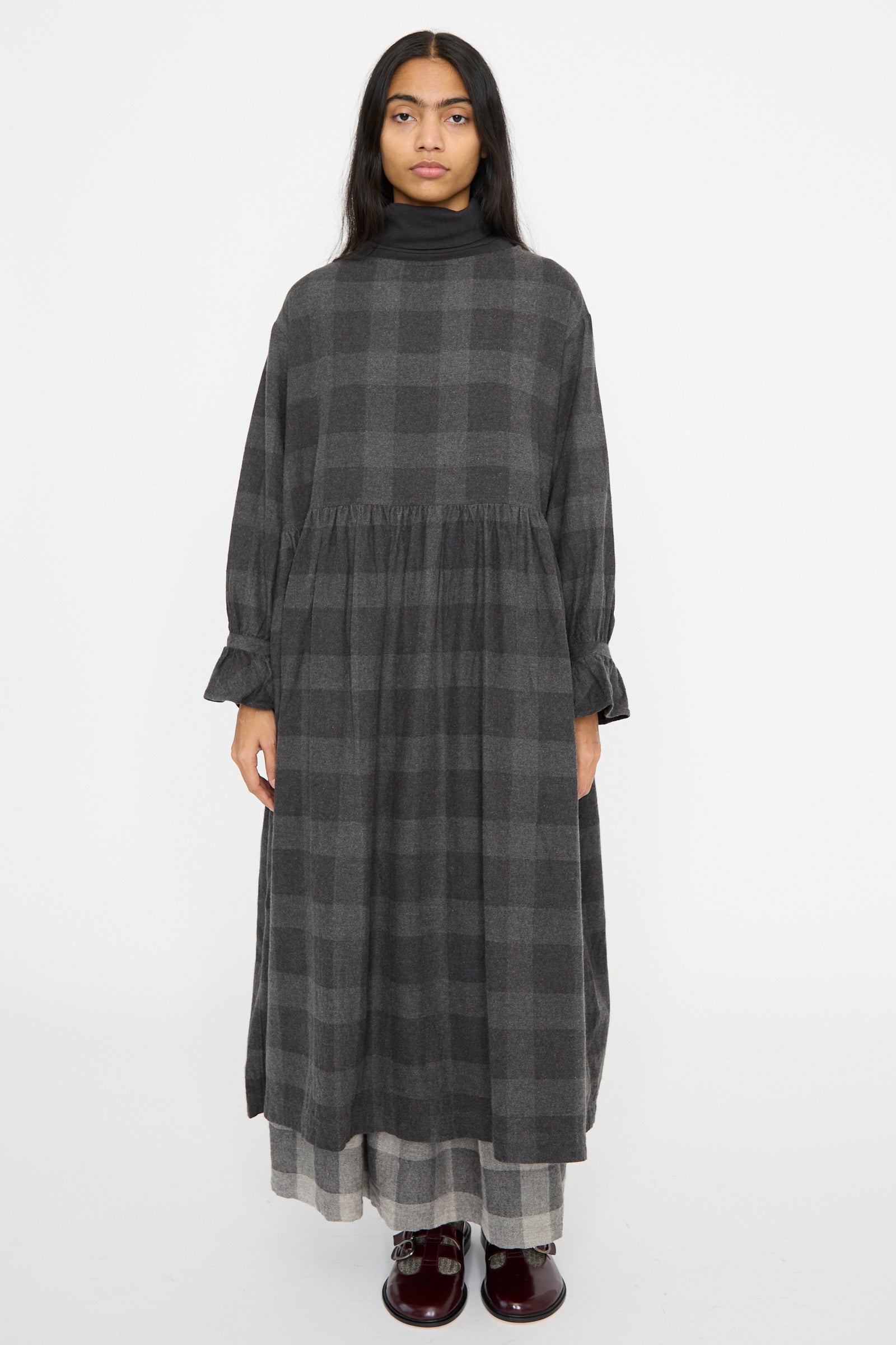 Wearing an Ichi Antiquités Block Check Flannel Dress in Charcoal with ruffled cuffs, a person stands against a plain white background, paired with burgundy shoes.