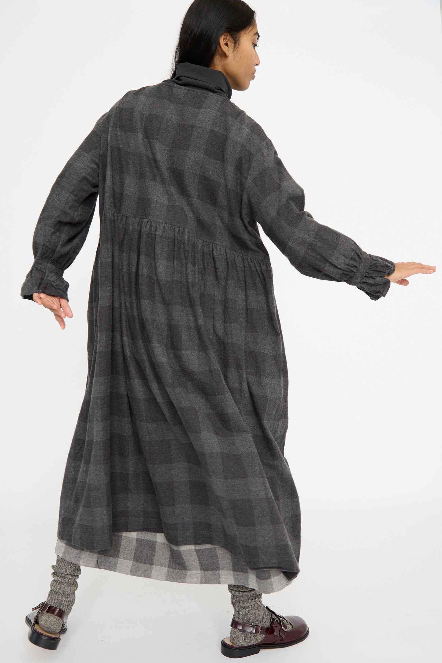 A person faces away n a Block Check Flannel Dress by Ichi Antiquités, showcasing a charcoal plaid design with ruffled cuffs, paired with dark shoes and posing against a plain white background.