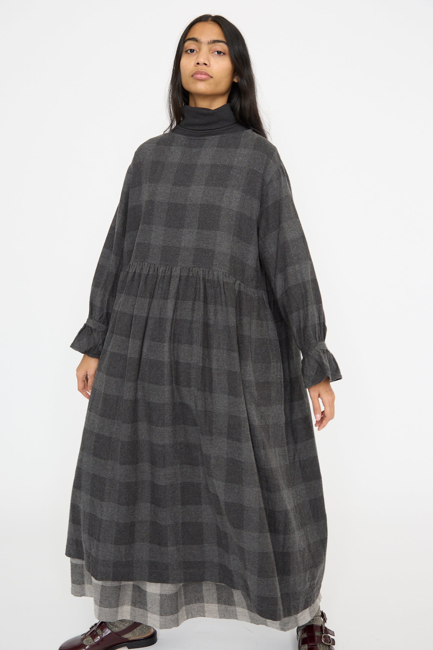 A person in the Ichi Antiquités Block Check Flannel Dress in Charcoal, featuring ruffled cuffs, and dark shoes stands against a plain white background.