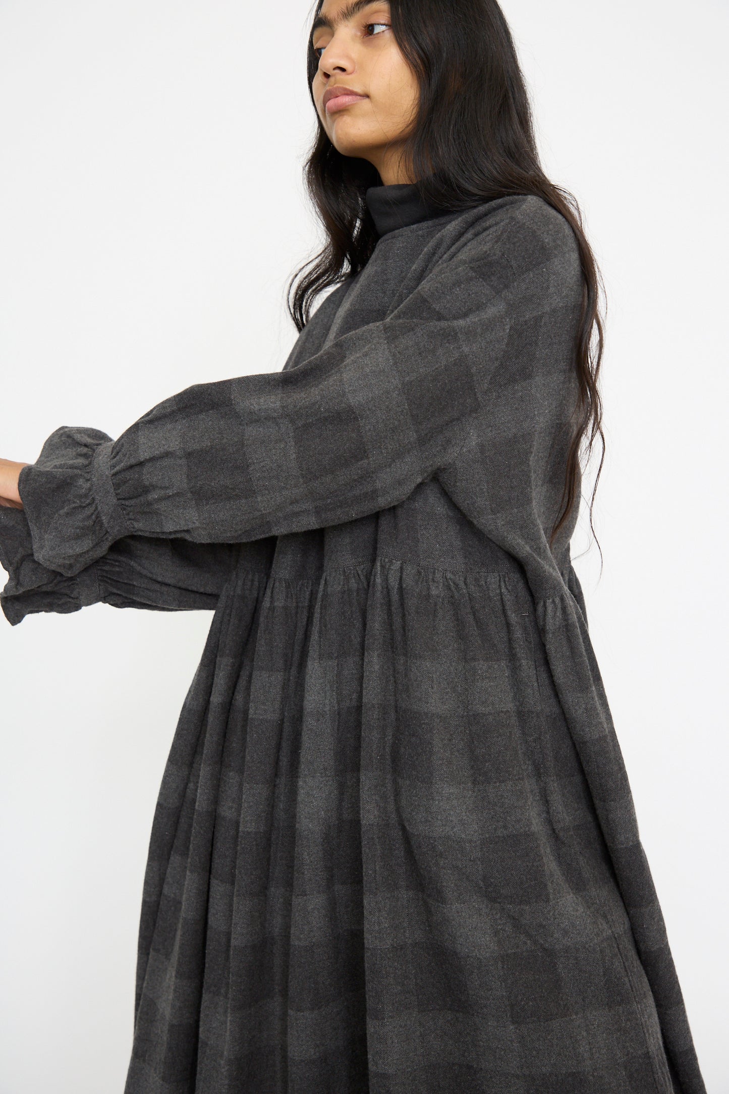 A person showcases the elegance of the Block Check Flannel Dress in Charcoal by Ichi Antiquités, featuring a long-sleeved design with ruffled cuffs against a plain background, highlighting its cotton/wool blend fabric.