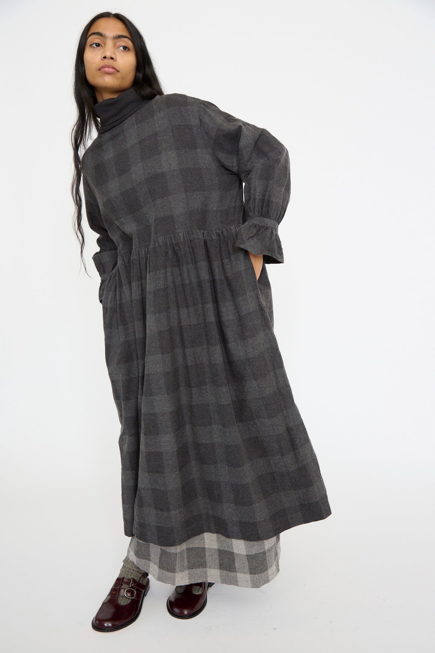 A model poses in the Block Check Flannel Dress by Ichi Antiquités, showcasing its dark plaid pattern, ruffled cuffs, and handy pockets, against a plain white background.