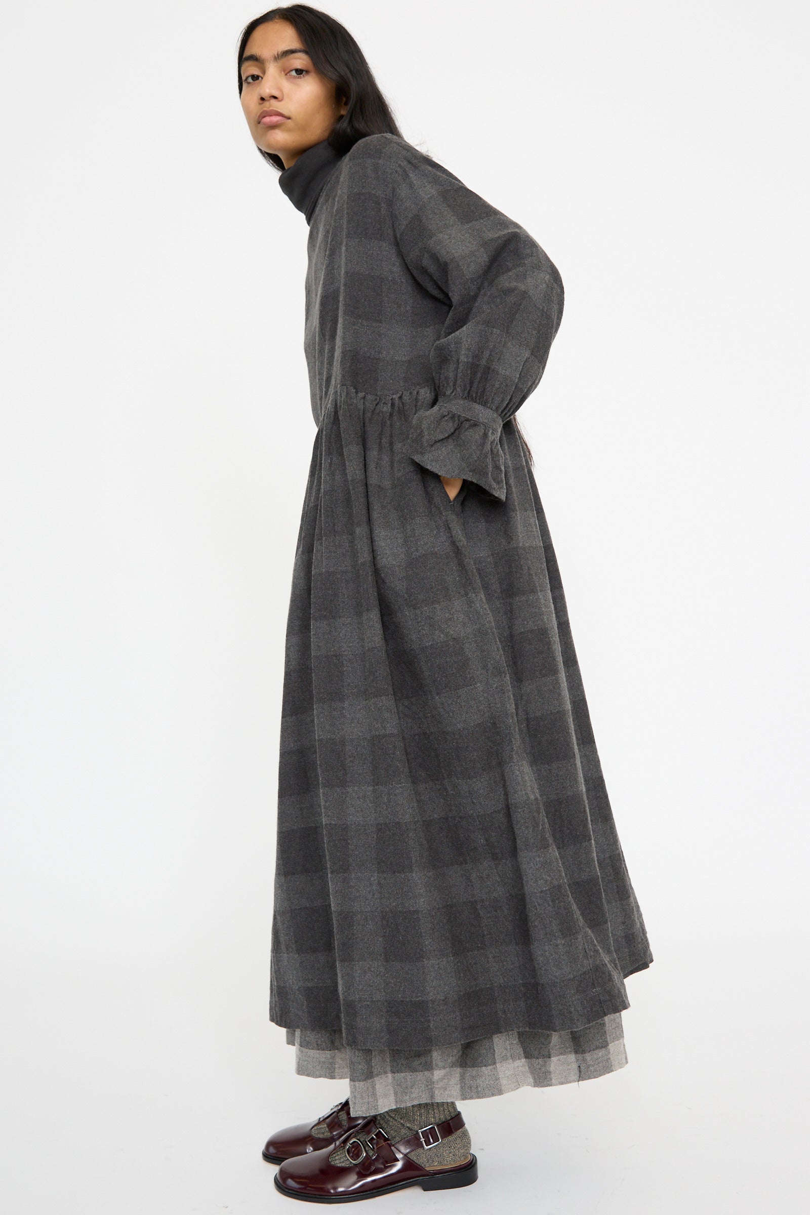 A person in a Block Check Flannel Dress by Ichi Antiquités, featuring a gray plaid design with ruffled cuffs, stands against a white background, facing sideways.