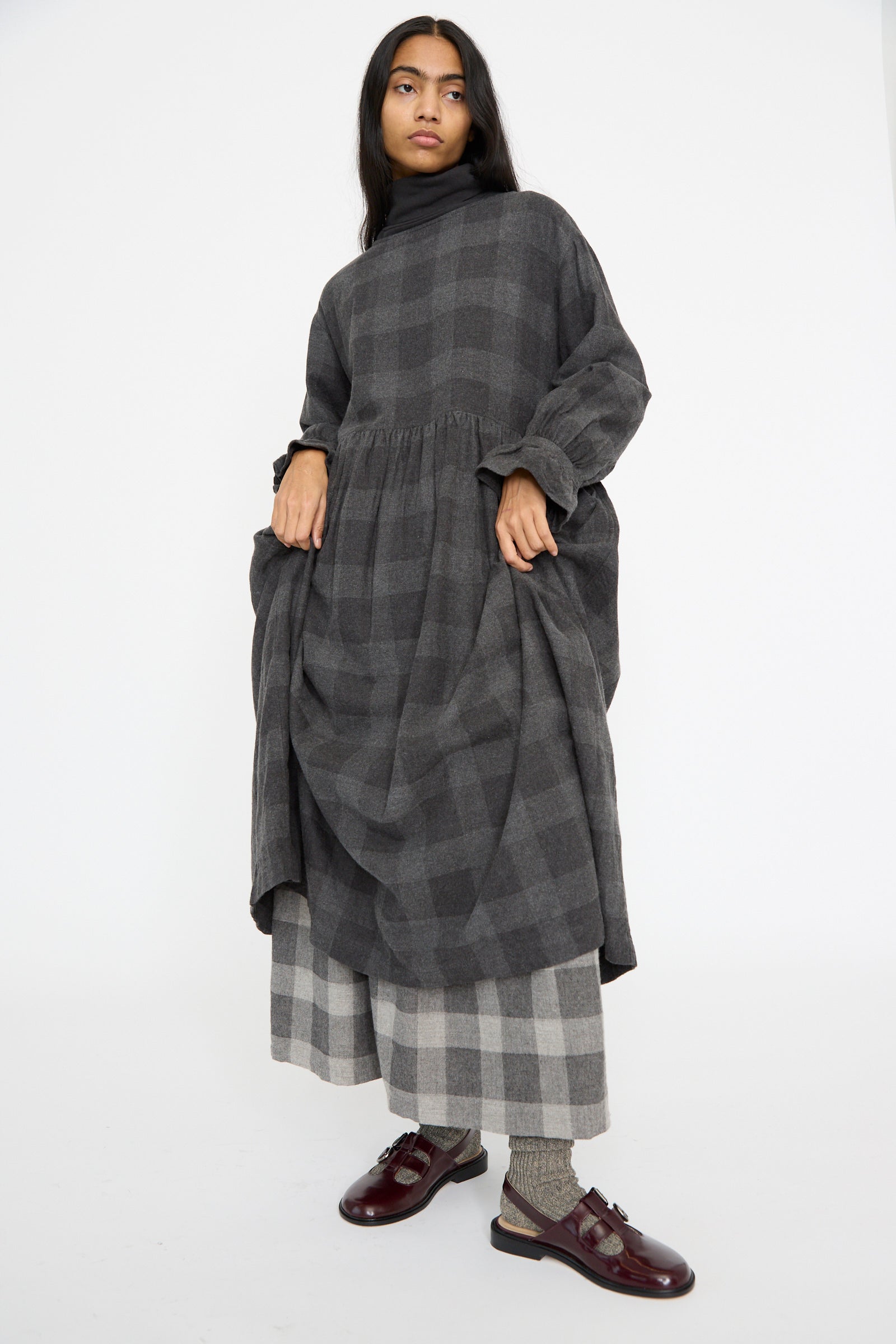 On a white backdrop, a person models the Block Check Flannel Dress in Charcoal by Ichi Antiquités, a maxi dress with long sleeves and a layered plaid design in soft cotton/wool blend, paired with dark shoes.