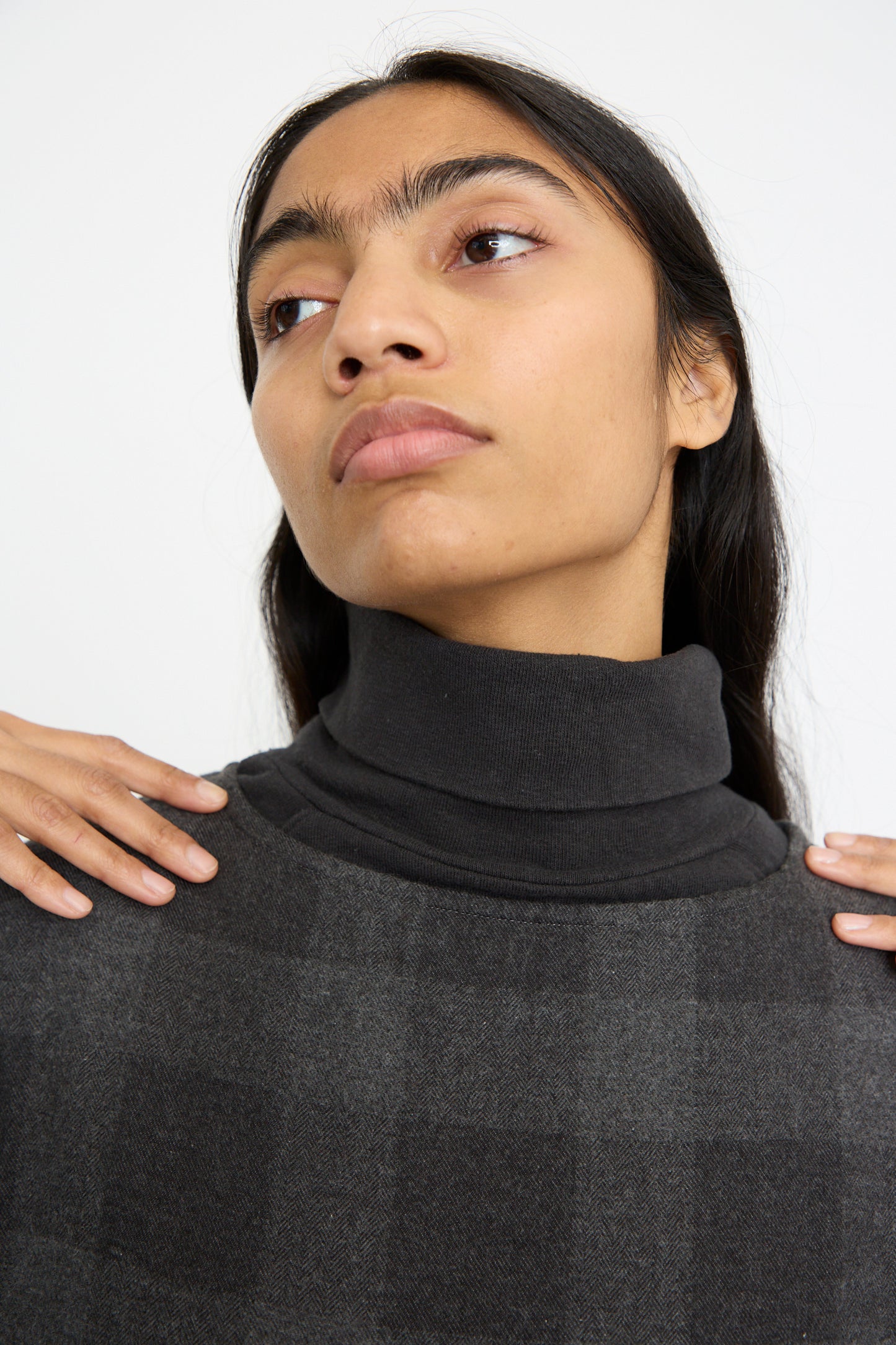 A person with long hair and a neutral expression wears the Block Check Flannel Dress in Charcoal by Ichi Antiquités, lightly touching their shoulders while looking slightly upward.