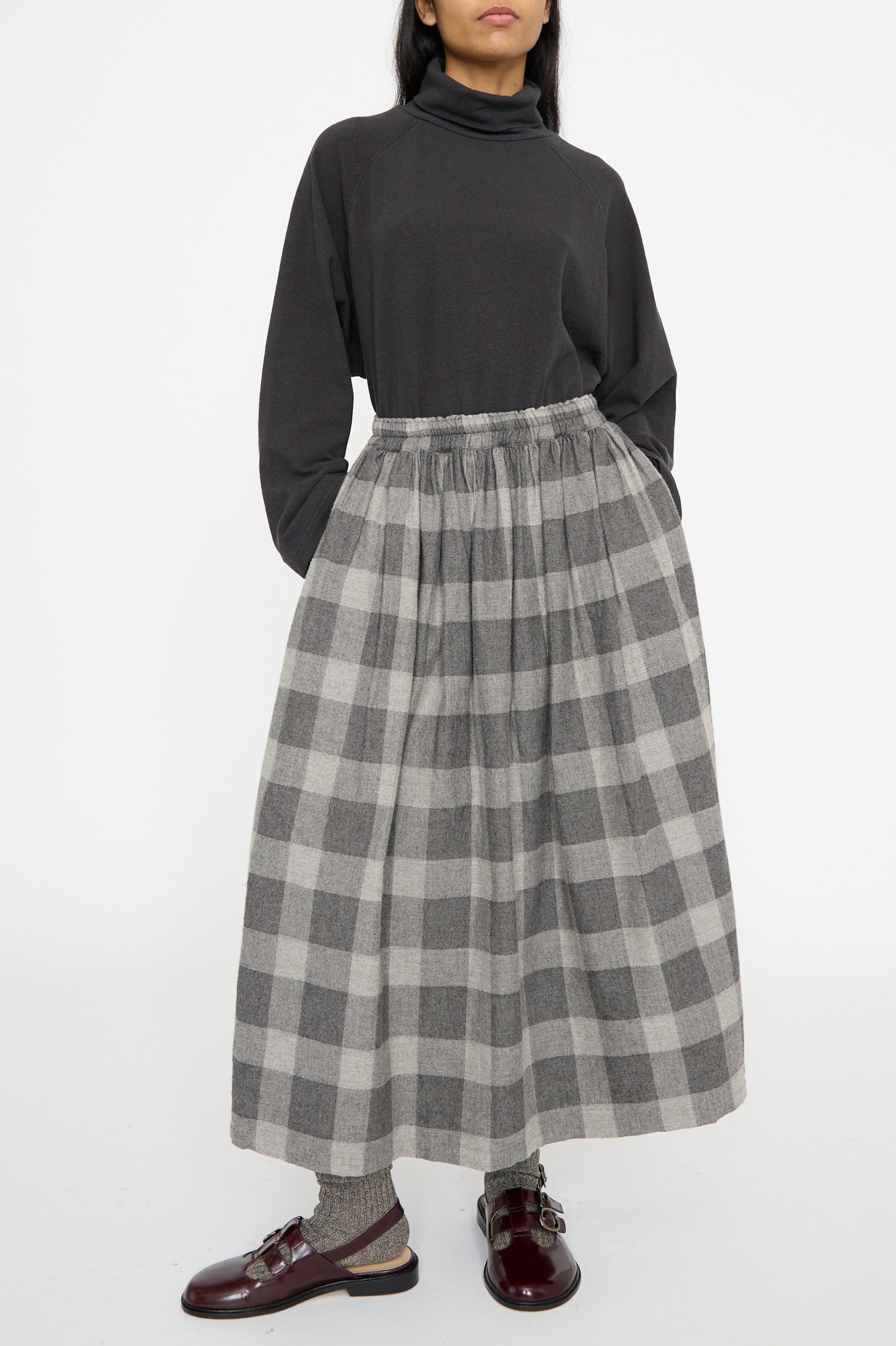 Against a white backdrop, the model wears a dark turtleneck and the Block Check Flannel Skirt in Gray by Ichi Antiquités, styled with dark socks and burgundy shoes.