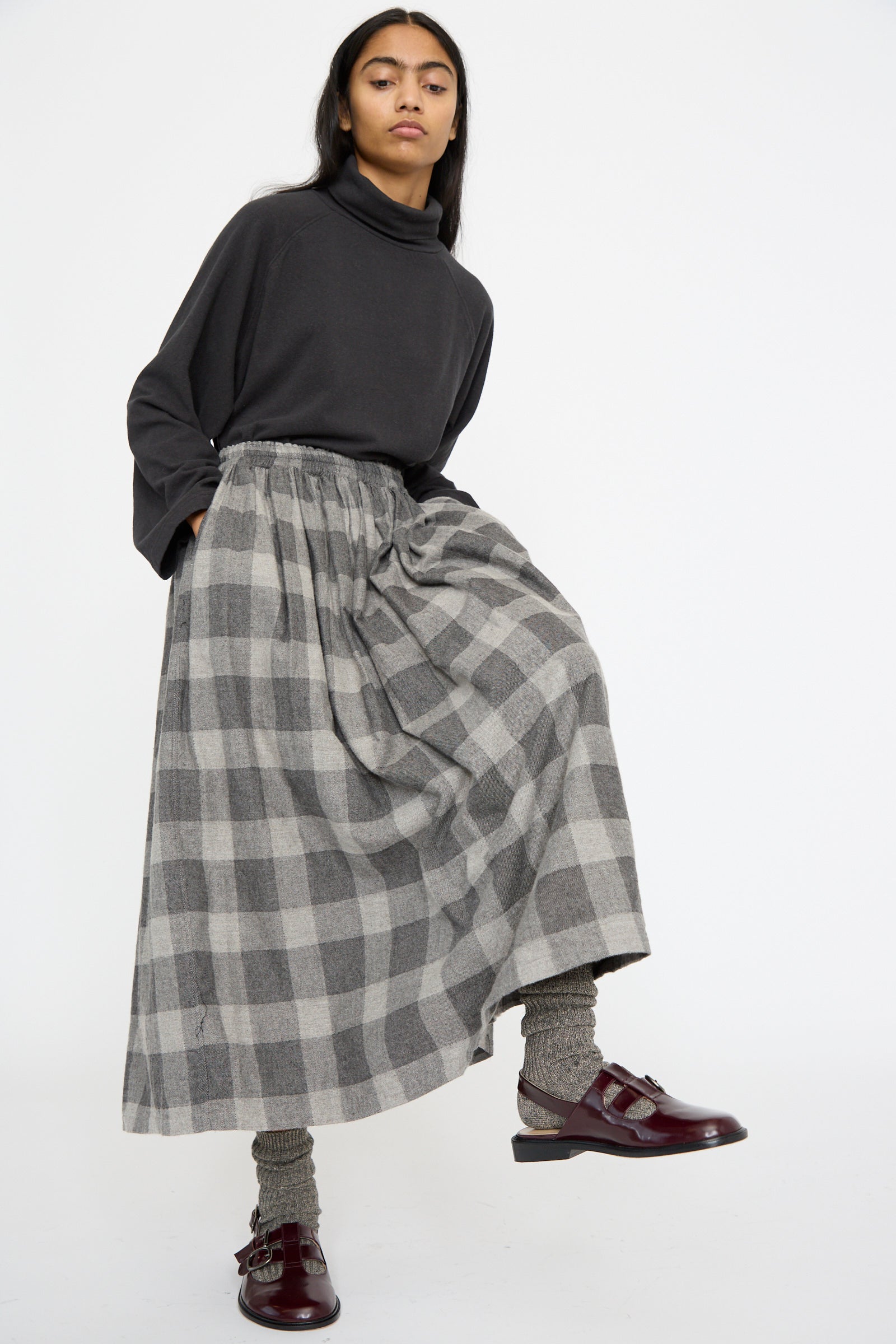 A person in an Ichi Antiquités Block Check Flannel Skirt in Gray, paired with a gray turtleneck, gray socks, and burgundy shoes, stands with one knee slightly bent.