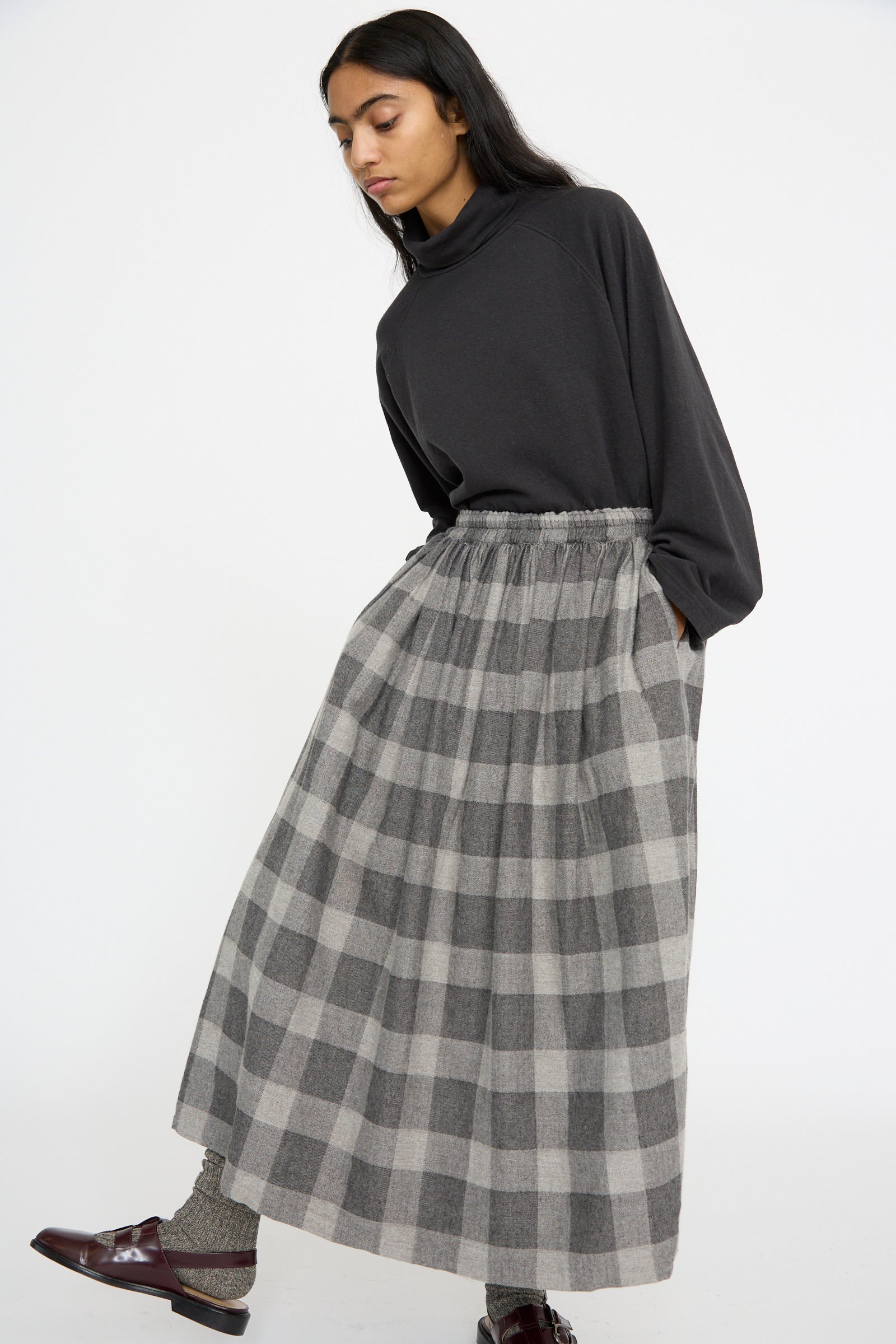 A person in a dark top and the Ichi Antiquités Block Check Flannel Skirt in Gray stands with one foot forward against a light background.