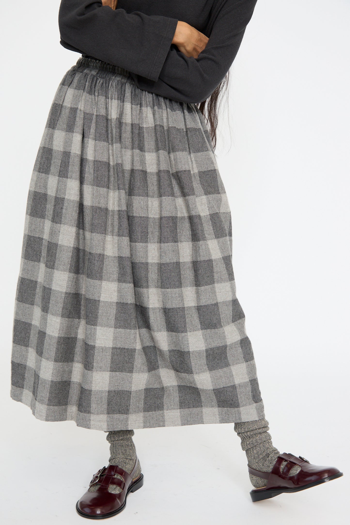 A person stands against a plain background wearing the Ichi Antiquités Block Check Flannel Skirt in Gray, a black top, and burgundy shoes with gray socks, seen below the waist. 