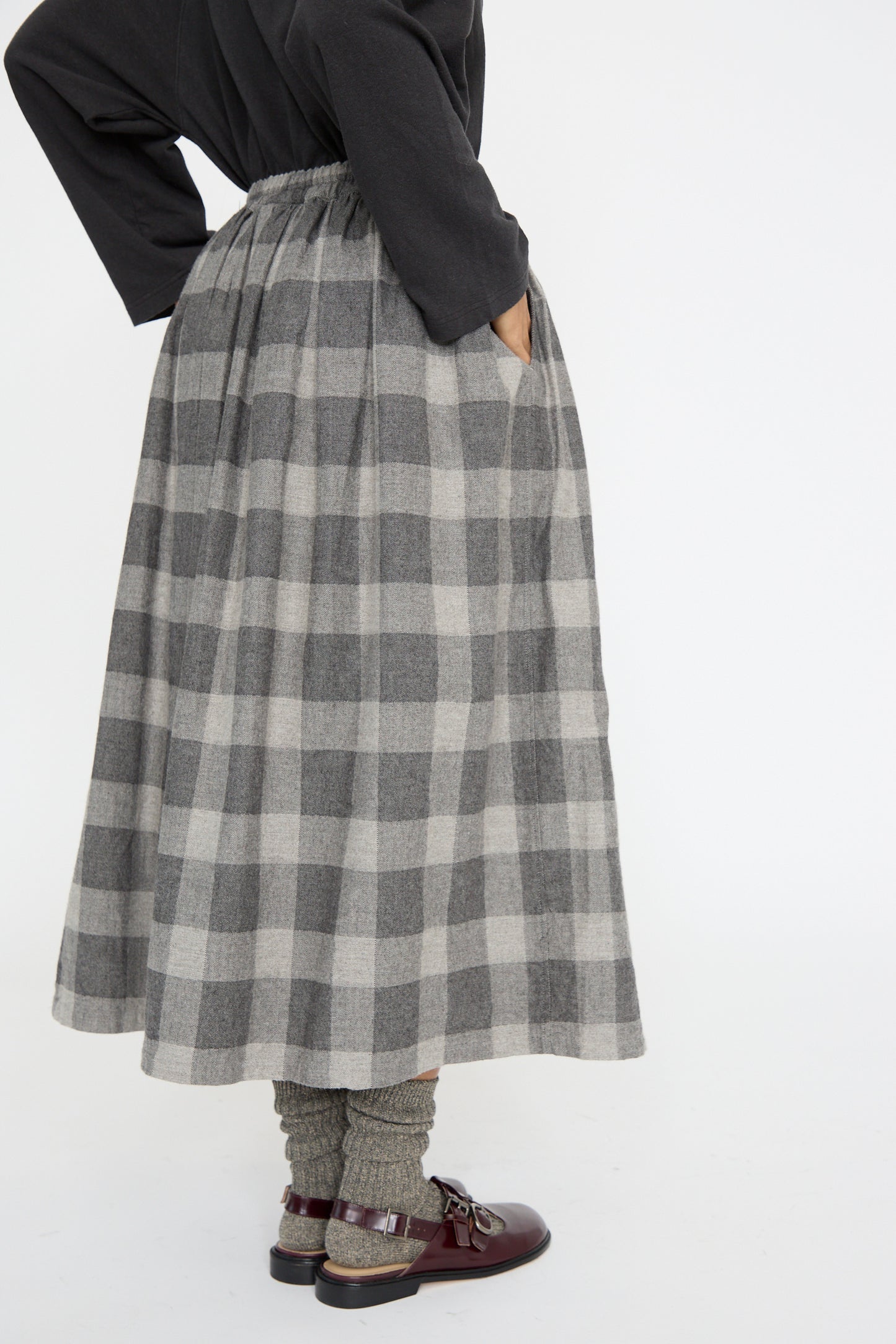 A person wearing a black top, Block Check Flannel Skirt in Gray by Ichi Antiquités, matching socks, and brown shoes stands with hands in skirt pockets against a plain background.