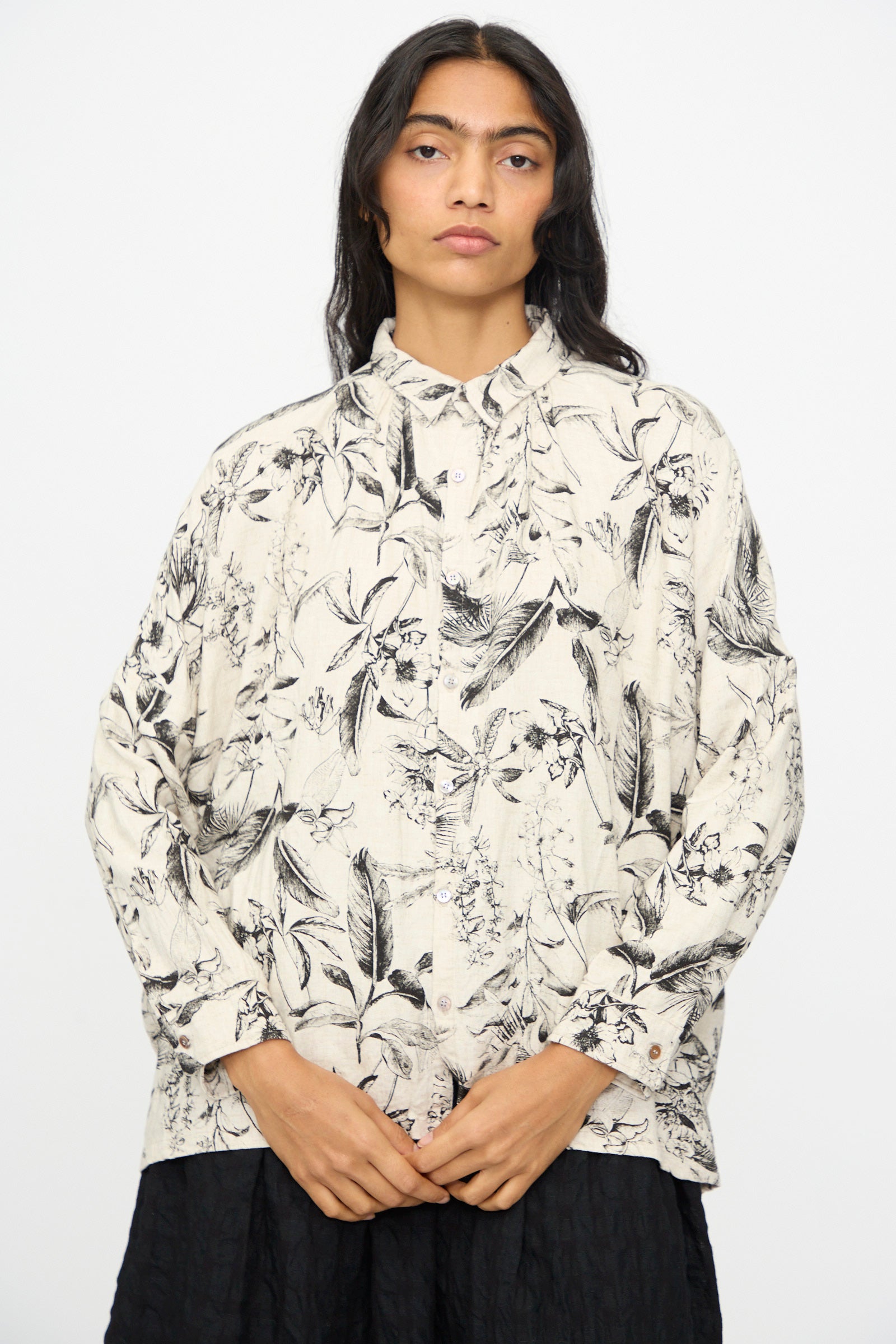 A person stands against a plain background wearing the Ichi Antiquités Botanical Print Shirt in Natural, a relaxed fit light-colored long sleeve shirt with a black floral pattern, paired with a black skirt.