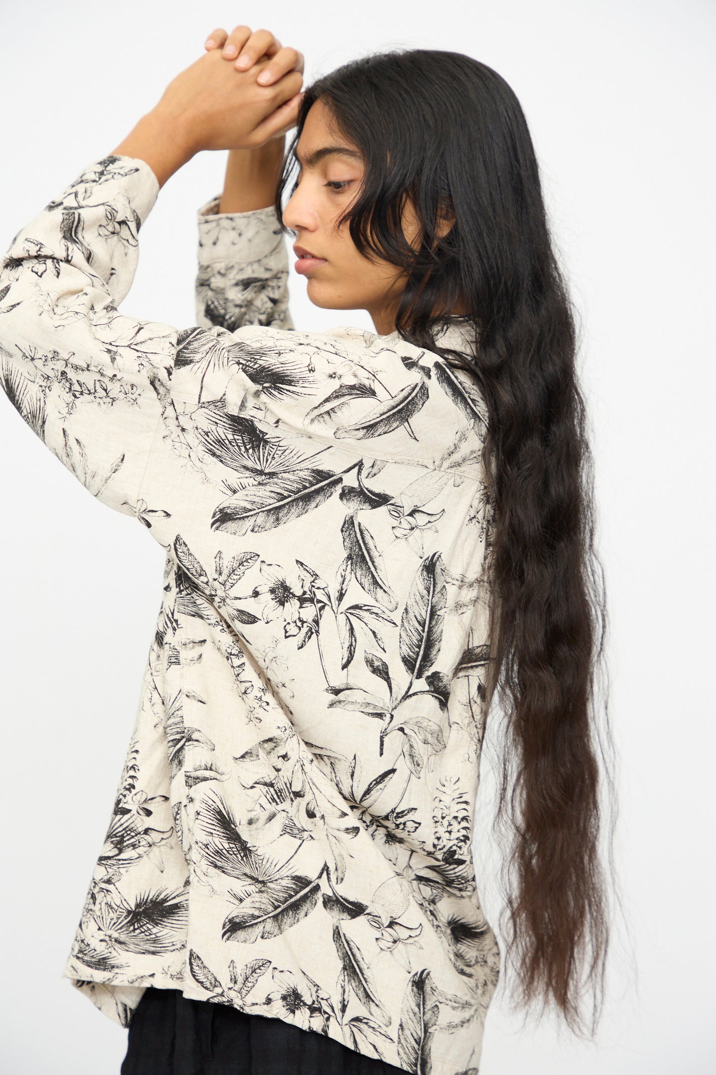 A person with long, wavy dark hair stands in profile against a plain light background, wearing the Ichi Antiquités Botanical Print Shirt in Natural with relaxed fit and long sleeves, hands clasped above their head.