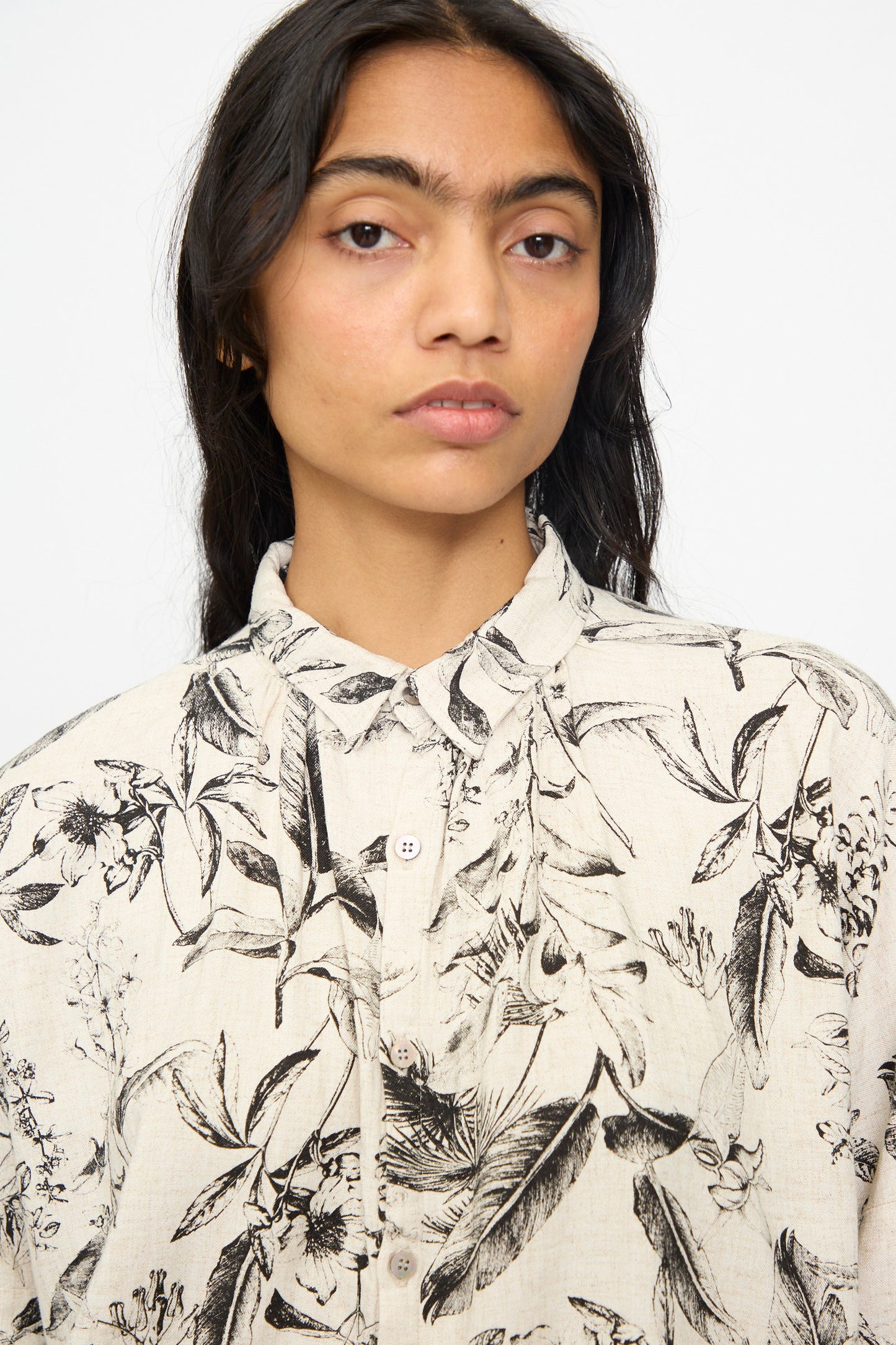 A person with long dark hair wears the Ichi Antiquités Botanical Print Shirt in Natural, a relaxed-fit beige linen button-up with a black botanical pattern, standing against a plain light background.