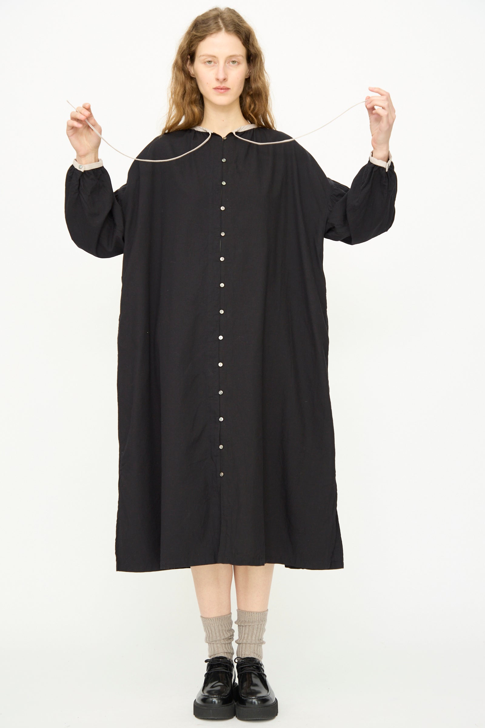 A person stands facing forward, wearing the Ichi Antiquités Cotton Cashmere Viyella Dress in Black, paired with beige socks and black shoes while holding a thin string with both hands.