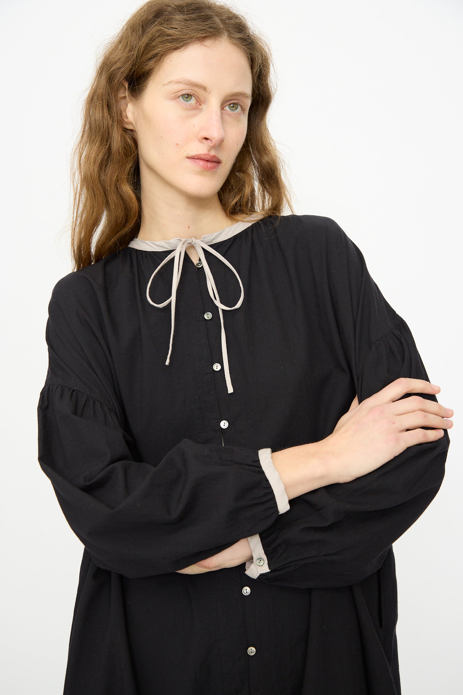 A person with long brown hair wears the Ichi Antiquités Cotton Cashmere Viyella Dress in Black, featuring a loose fit with light trim and a bow at the collar, standing against a plain white background.