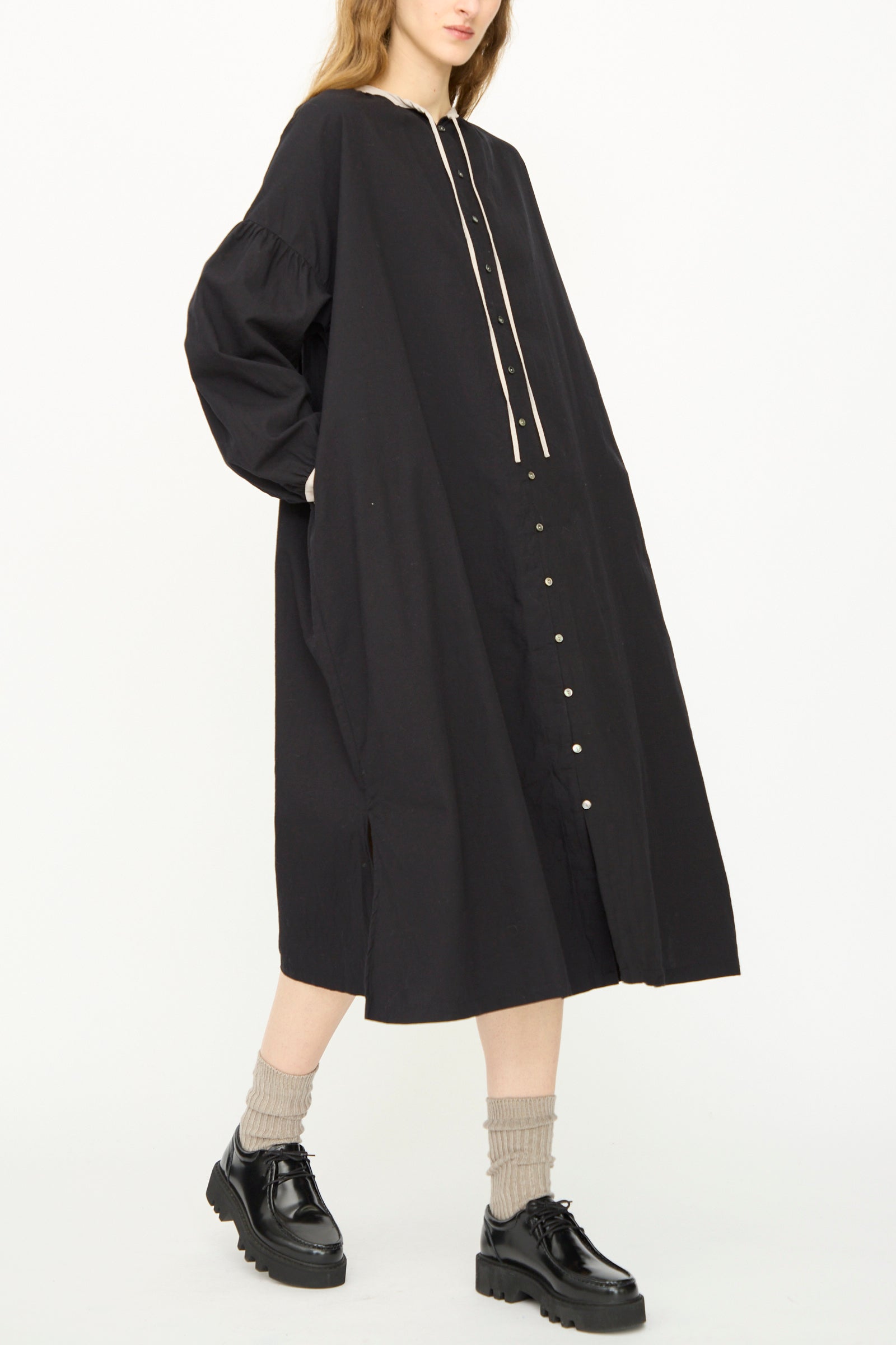 A person wears the Ichi Antiquités Cotton Cashmere Viyella Dress in Black—loose-fitting with long sleeves—paired with chunky black shoes and beige socks, standing against a plain white background.