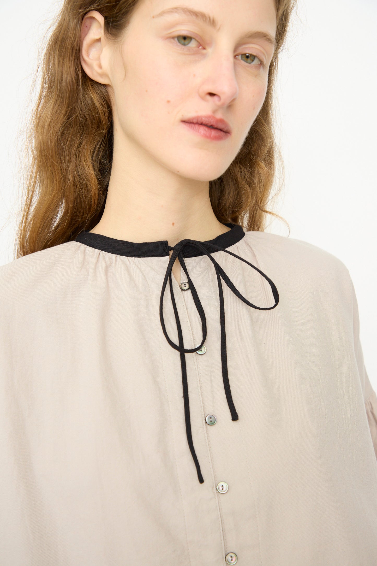 A person with long light brown hair wears the Ichi Antiquités Cotton Cashmere Viyella Shirt in Natural, featuring a black collar and tie detail, gazing slightly to the side against a plain background.