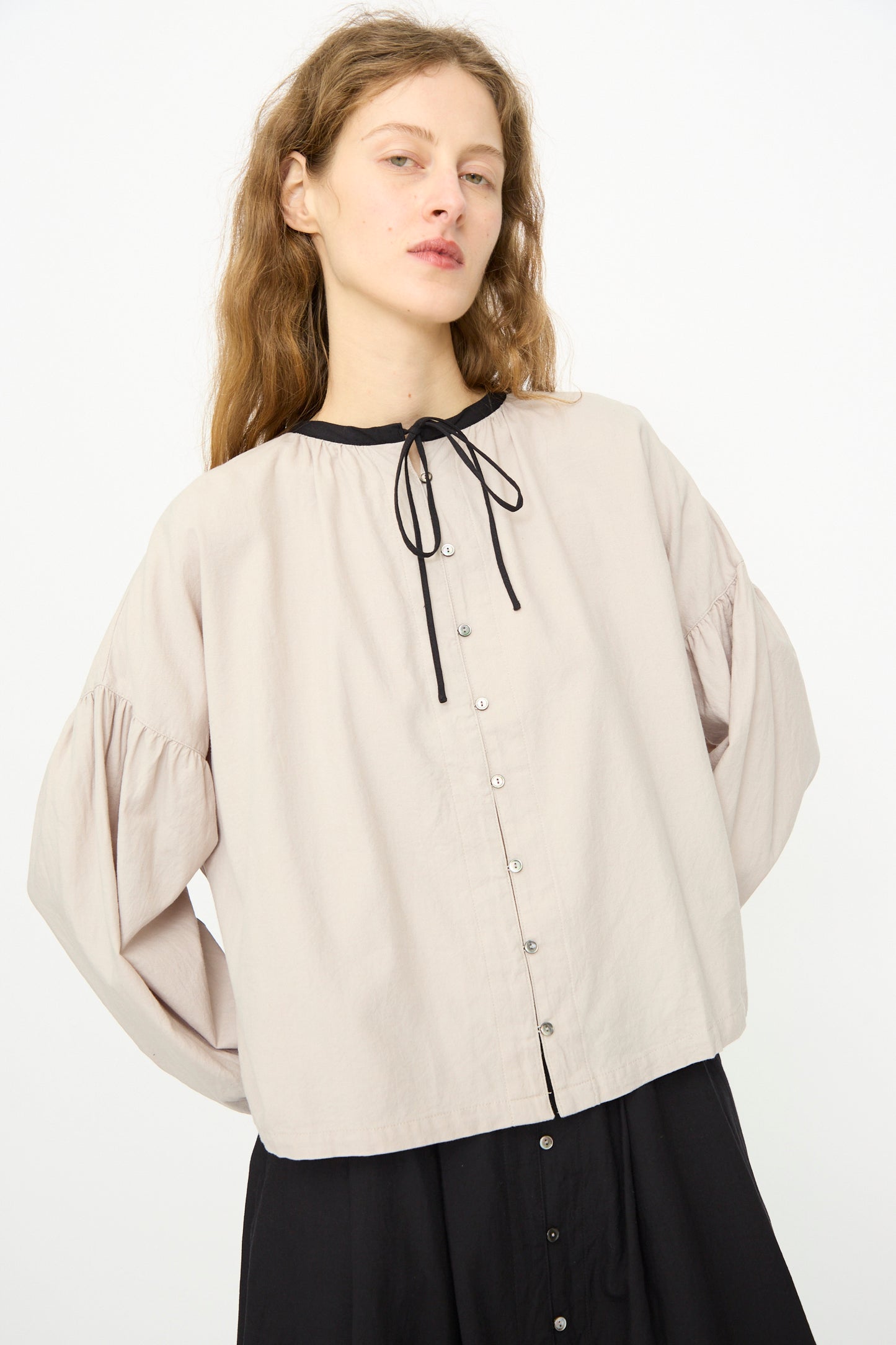 A person with long light brown hair wears the Ichi Antiquités Cotton Cashmere Viyella Shirt in Natural, featuring puffy sleeves and a black neck ribbon, paired with a black skirt, standing against a plain white background.