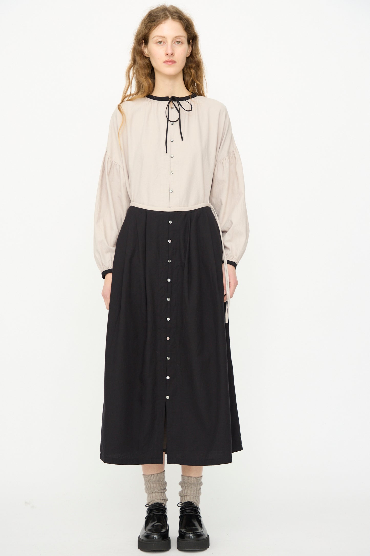 A woman faces forward wearing a beige long-sleeve blouse with a black tie at the neck, the Ichi Antiquités Cotton Cashmere Viyella Skirt in Black (made in Japan), paired with black shoes and beige socks.