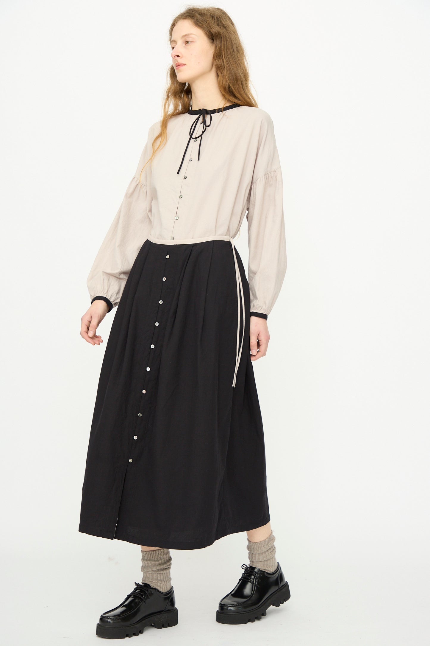 A person wears a long-sleeve beige blouse with a black tie at the neck, tan socks, black platform shoes, and the Cotton Cashmere Viyella Skirt in Black by Ichi Antiquités. 