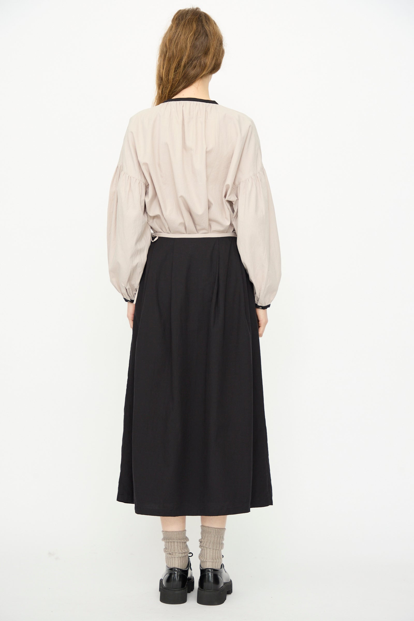 A person stands facing away against a white background, wearing a beige long-sleeve blouse, gray socks, black shoes, and the Ichi Antiquités Cotton Cashmere Viyella Skirt in Black.