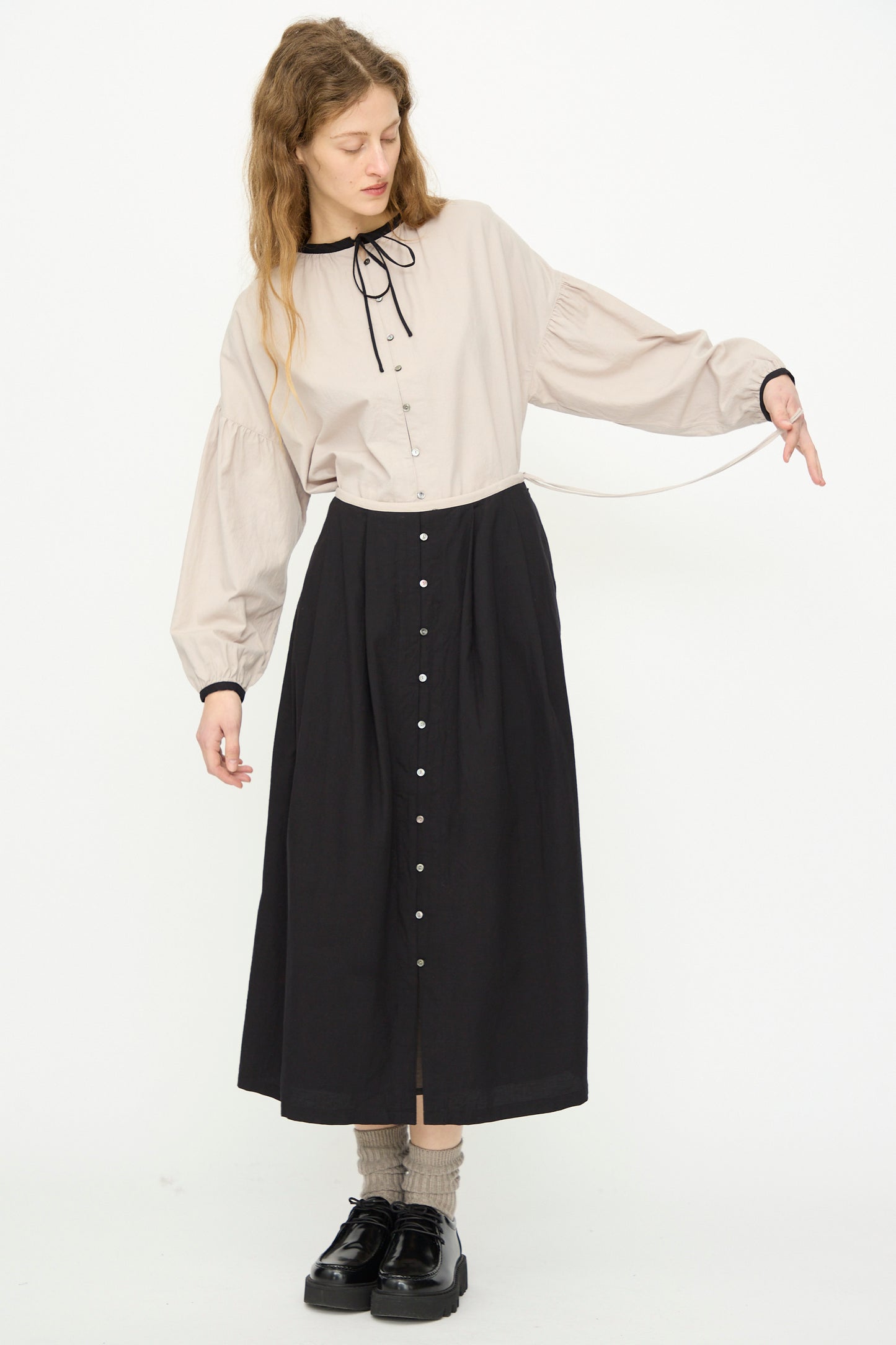 A woman stands against a white background wearing a beige long-sleeve blouse with a black ribbon, gray socks, black shoes, and the Ichi Antiquités Cotton Cashmere Viyella Skirt in Black.