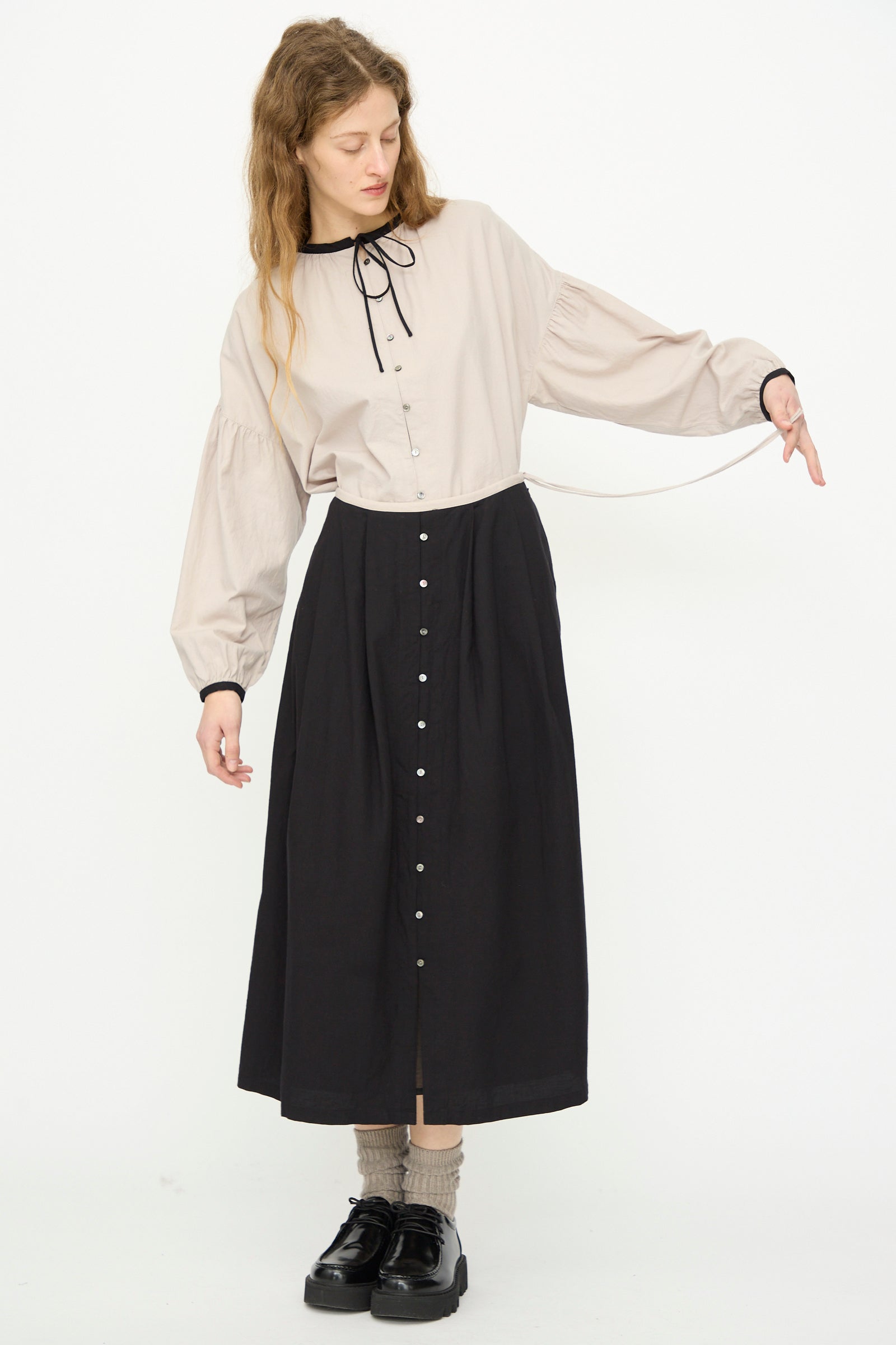 A woman stands against a white background wearing a beige long-sleeve blouse with a black ribbon, gray socks, black shoes, and the Ichi Antiquités Cotton Cashmere Viyella Skirt in Black.